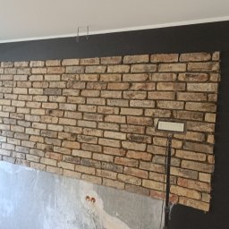 Kam.gru.bud. - Ściana z cegieł klinkierowych w trakcie montażu, widoczne fragmenty surowej ściany poniżej, oświetlenie halogenowe i czarna ściana w tle.
