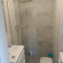 Wykończona łazienka z kabiną prysznicową typu walk-in, umywalką z szafką, szarą marmurkową glazurą na ścianach i podłodze, oraz detalami jak okrągły dywanik i miska z wodą i gąbką.