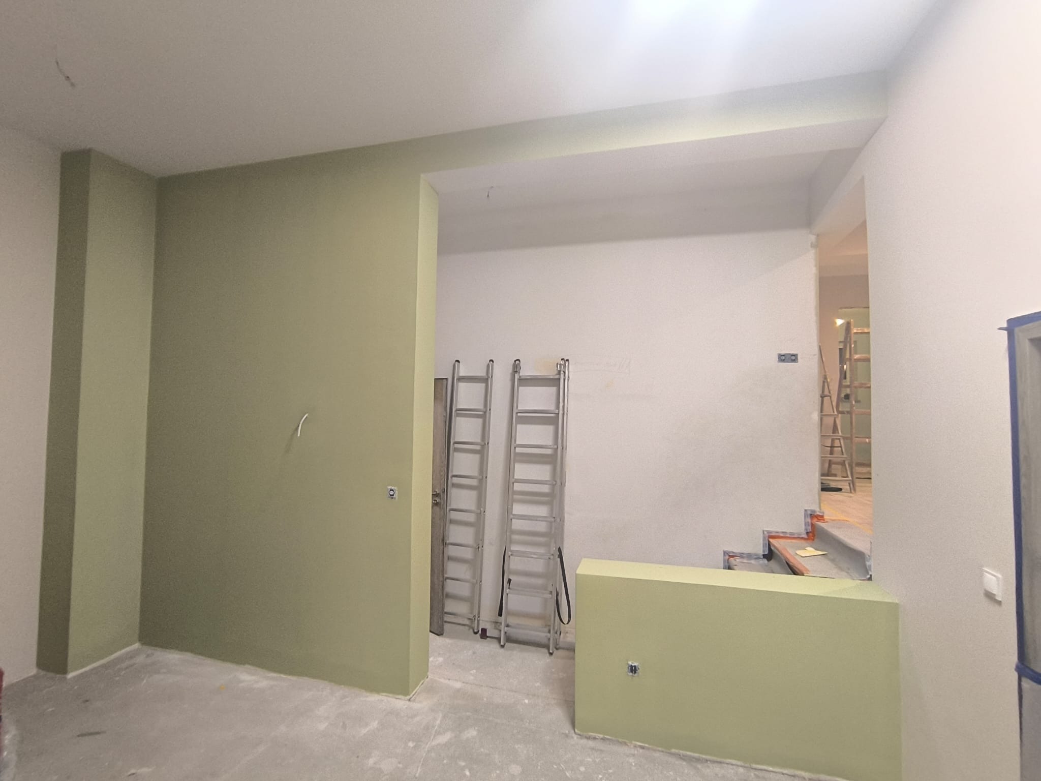Świeżo pomalowane na oliwkowy kolor wnętrze z dwiema aluminiowymi drabinami opartymi o ścianę. Widoczne gniazdka elektryczne i fragment schodów w tle.