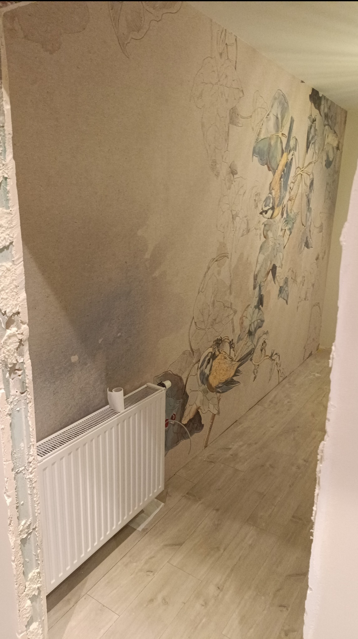 Ściana w trakcie malowania z motywem roślinnym, widoczny biały grzejnik i fragment podłogi z paneli, surowy stan wykończenia przy futrynie drzwi.