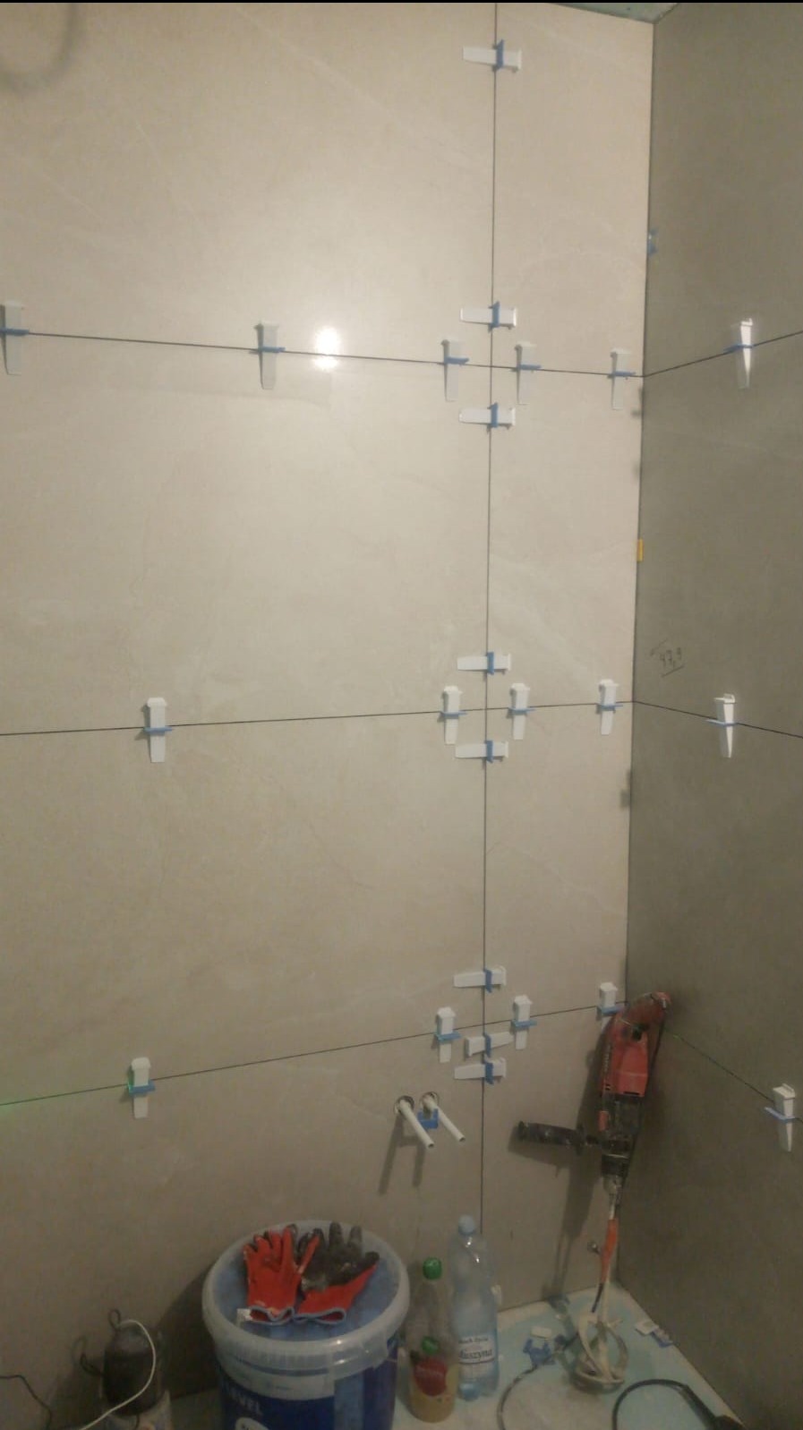 Ściana łazienki w trakcie układania płytek, widoczne krzyżyki dystansowe, poziomica laserowa, mieszadło do zaprawy, wiadro z rękawicami i butelki z wodą.