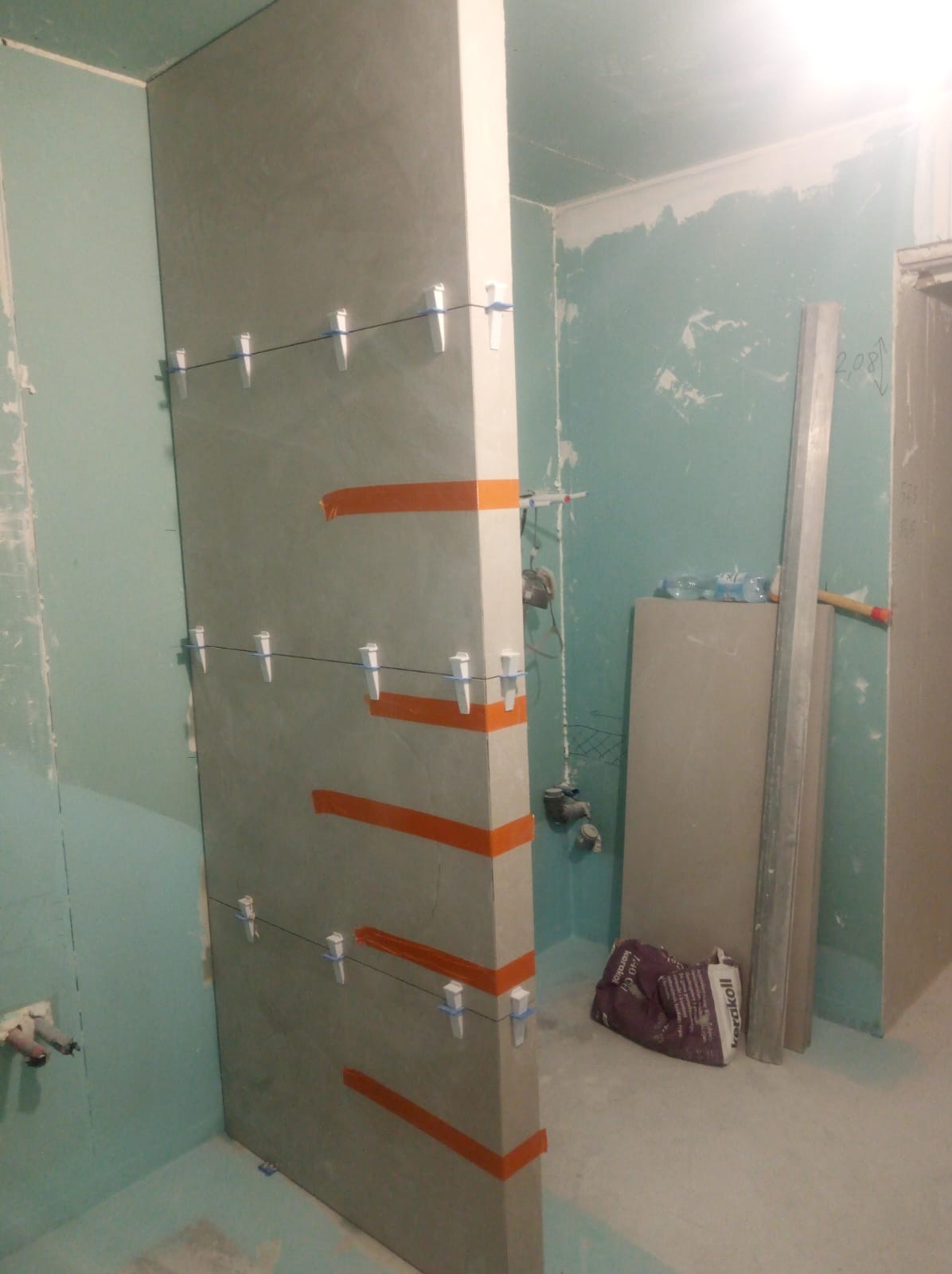 Płyta gipsowo-kartonowa mocowana do ściany w łazience za pomocą pomarańczowej taśmy i plastikowych klinów do poziomowania płytek, widoczny worek z zaprawą klejową Kerakoll.