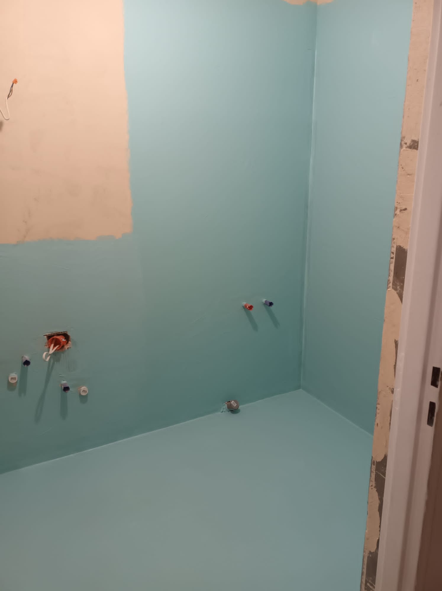 Świeżo pomalowana na turkusowo łazienka w trakcie prac wykończeniowych, widoczne wystające rury i kable, fragment ściany w kolorze beżowym z niedomalowanym obszarem, surowe obramowanie drzwi.