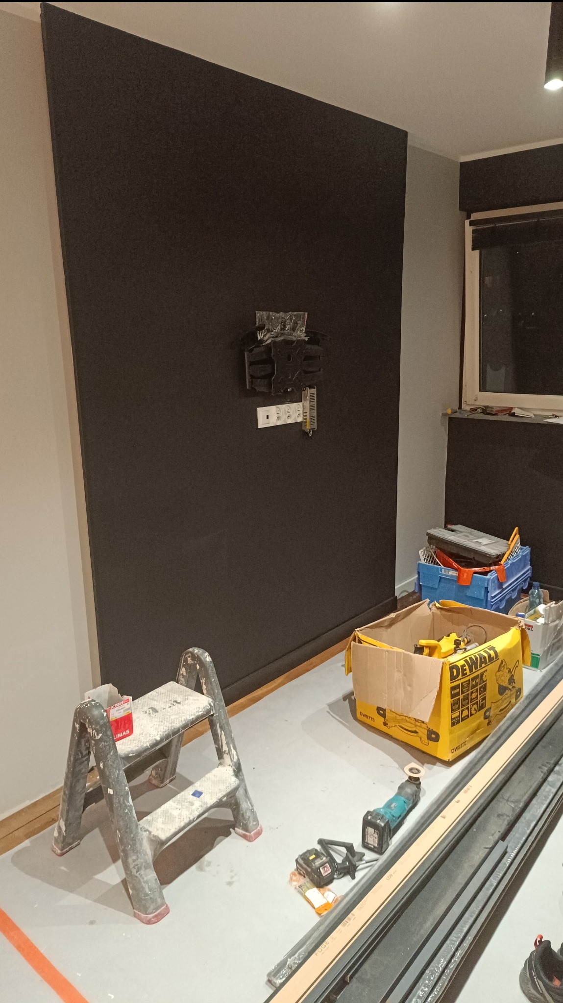 Przygotowanie do montażu telewizora na czarnej ścianie: widoczny uchwyt, gniazdka elektryczne, drabinka, narzędzia DeWalt w kartonie i listwy przypodłogowe na podłodze.