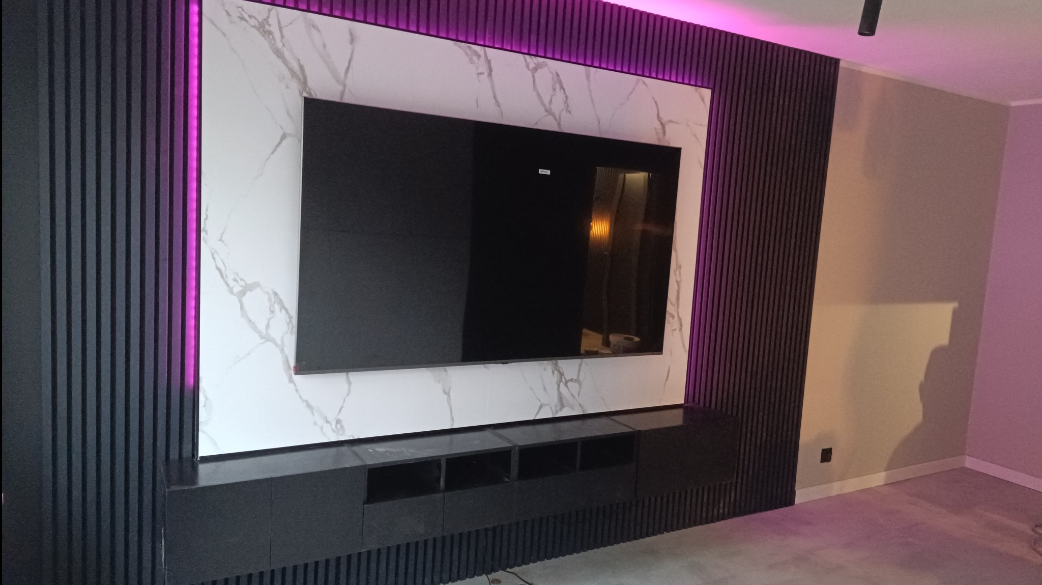 Nowoczesna ściana telewizyjna z podświetleniem LED, wykończona panelami z ciemnego drewna i marmurową płytą, z czarną szafką RTV na dole.