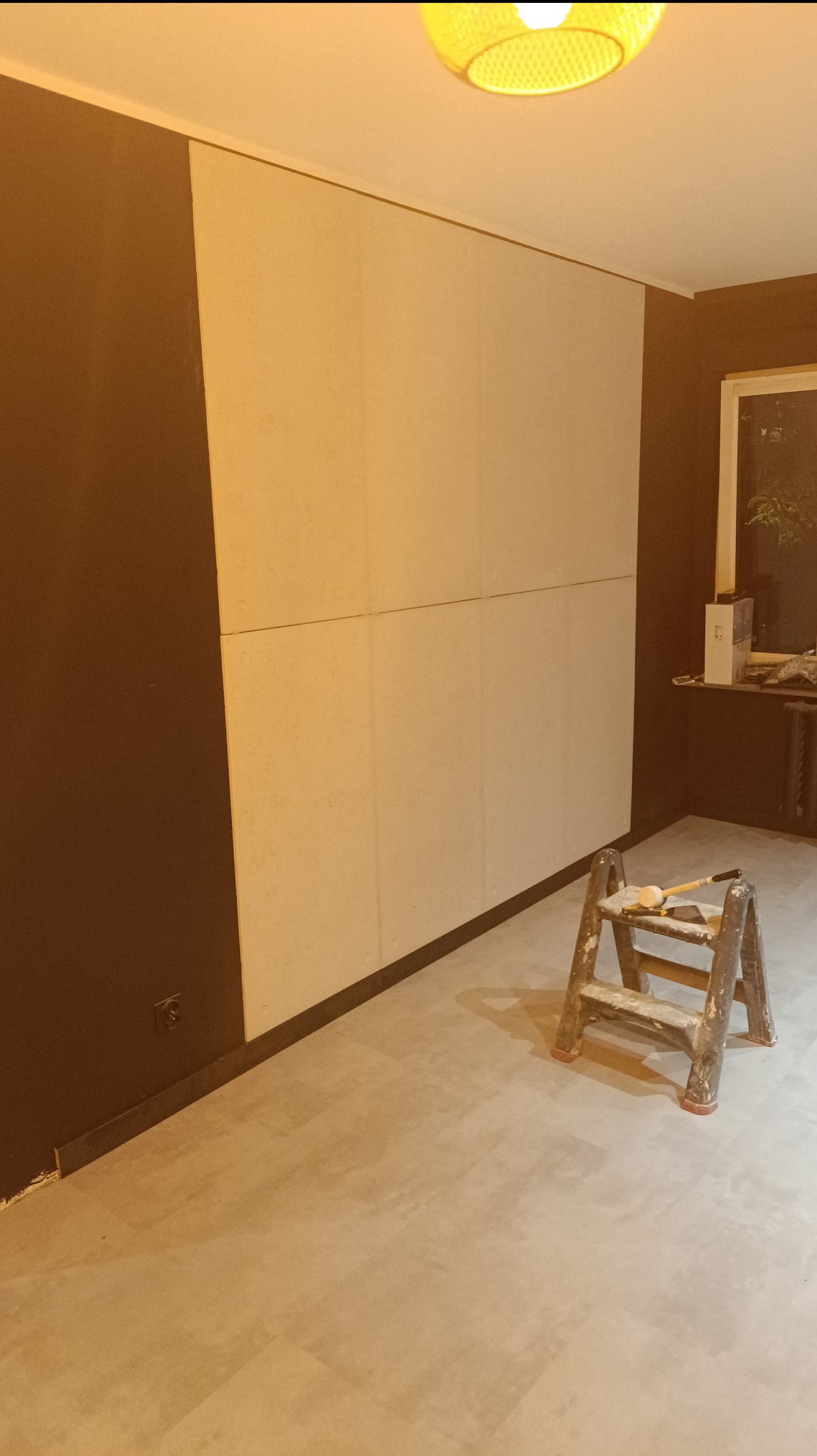 Ściana w trakcie remontu: fragment pomalowany na ciemny brąz, reszta w jasnych płytach, drabina z narzędziami malarskimi w pomieszczeniu z szarą podłogą imitującą beton.