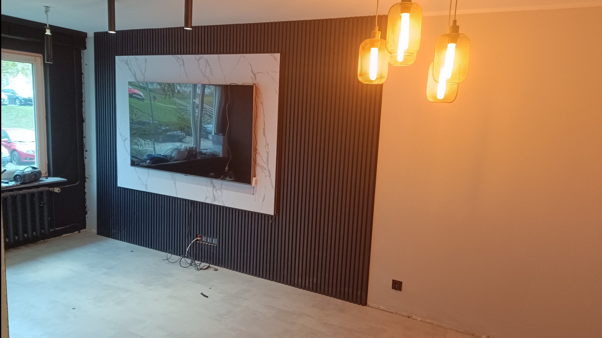 Salon po metamorfozie: ciemne, pionowe panele ścienne z telewizorem w marmurowej ramie, wiszące lampy z żarnikiem, grzejnik żeliwny pod oknem, widoczne kable elektryczne.
