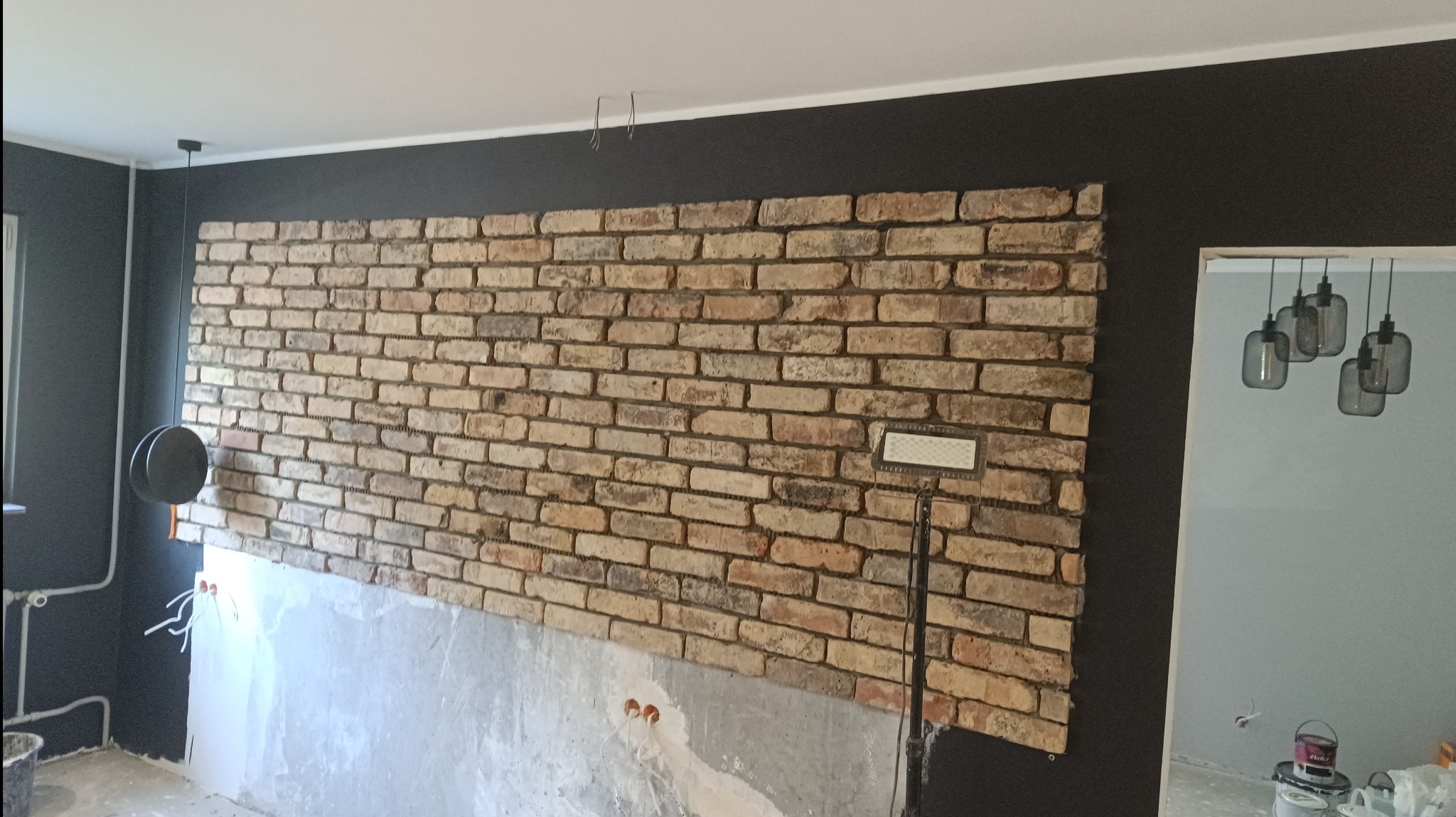 Ściana z cegieł klinkierowych w trakcie montażu, widoczne fragmenty surowej ściany poniżej, oświetlenie halogenowe i czarna ściana w tle.