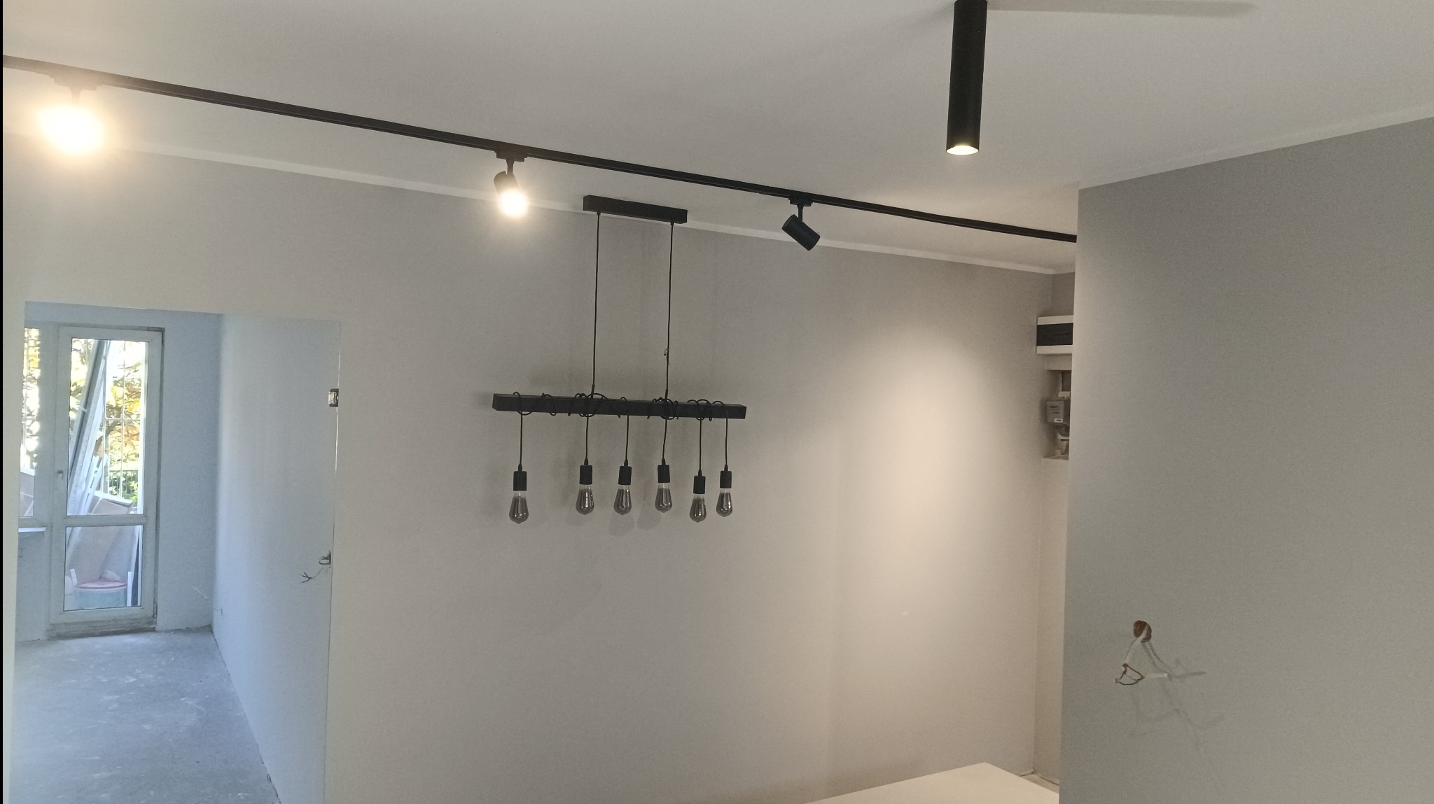 Wnętrze w trakcie wykończenia, widoczne szare ściany po szpachlowaniu, czarne szyny oświetleniowe z reflektorami i wisząca lampa z żarówkami Edisona, oraz wystające kable elektryczne.