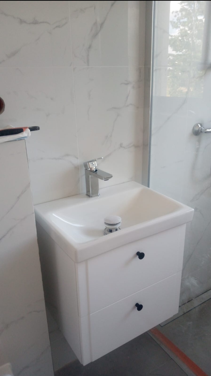 Nowoczesna biała umywalka z szafką z czarnymi uchwytami, chromowana bateria, marmurowe płytki ścienne i fragment kabiny prysznicowej.