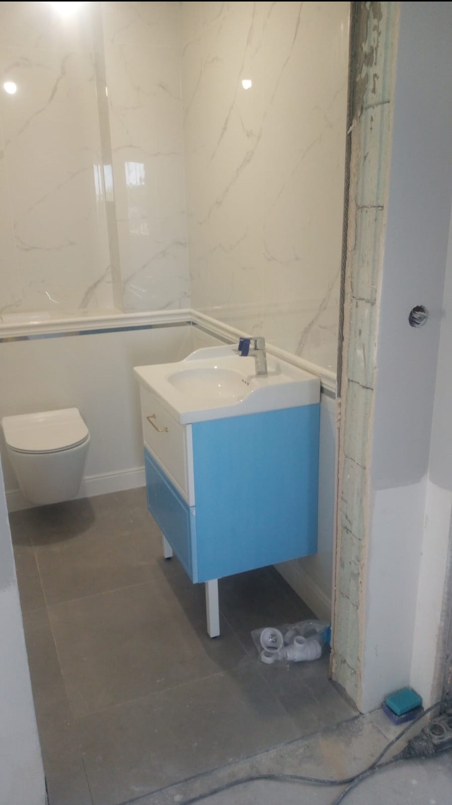 Wykończona łazienka z wiszącą toaletą i szafką umywalkową w kolorze błękitu, widoczne białe płytki imitujące marmur na ścianach i szare płytki na podłodze, w rogu przy futrynie leżą akcesoria...