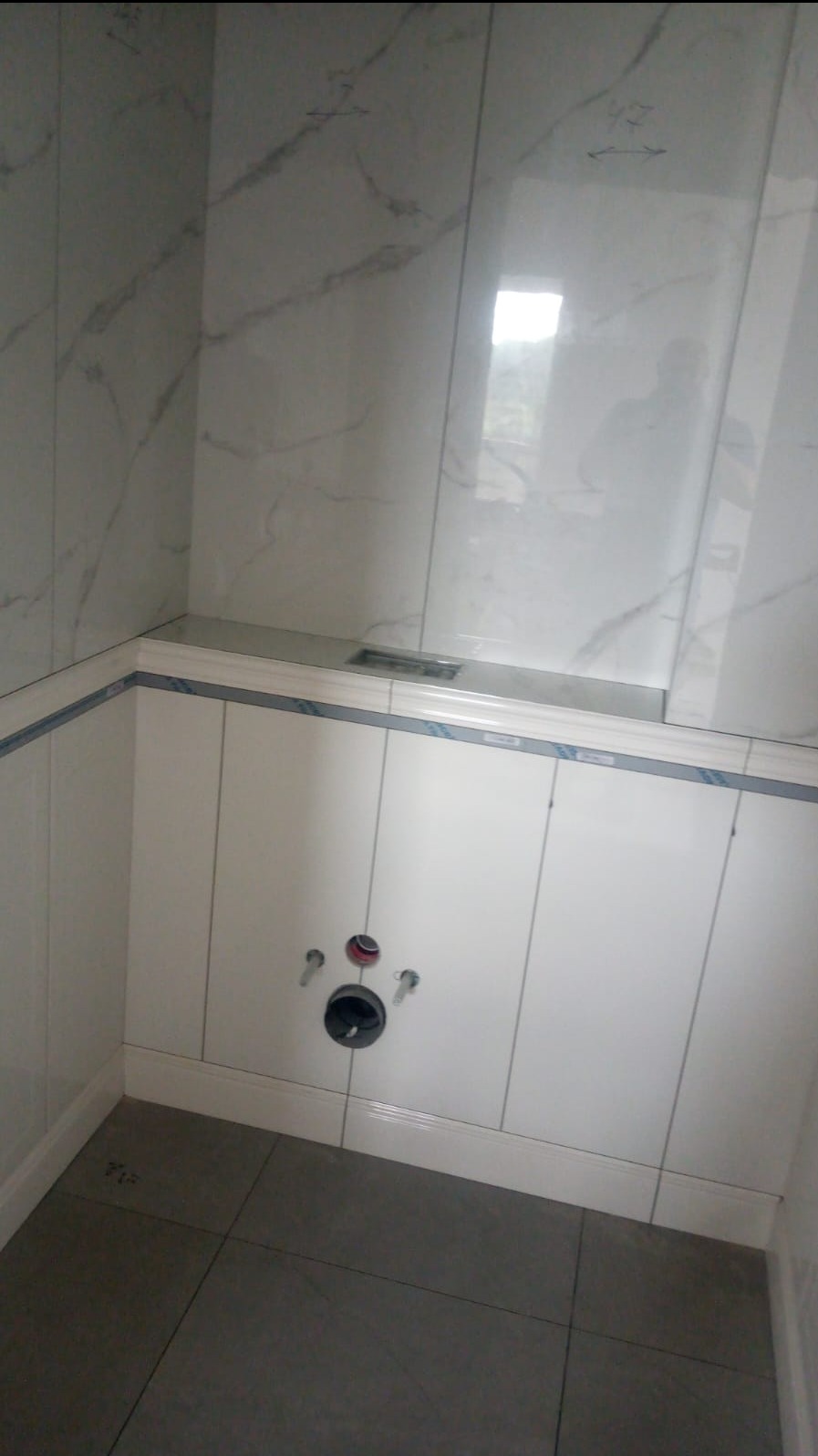 Łazienka w trakcie remontu: białe płytki imitujące marmur na ścianach, szare płytki podłogowe, widoczne przyłącza wodno-kanalizacyjne przed montażem sanitariatów.