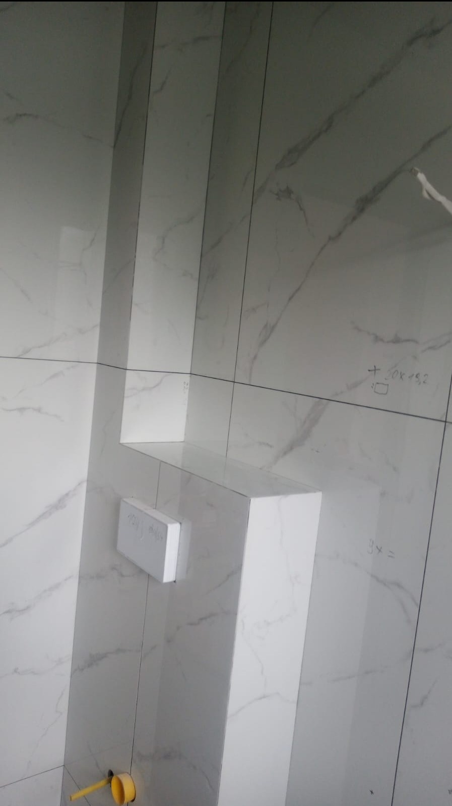 Ściana łazienki wyłożona białymi płytkami imitującymi marmur, z widoczną zabudową stelaża podtynkowego WC i fragmentem instalacji hydraulicznej w żółtym kolorze.