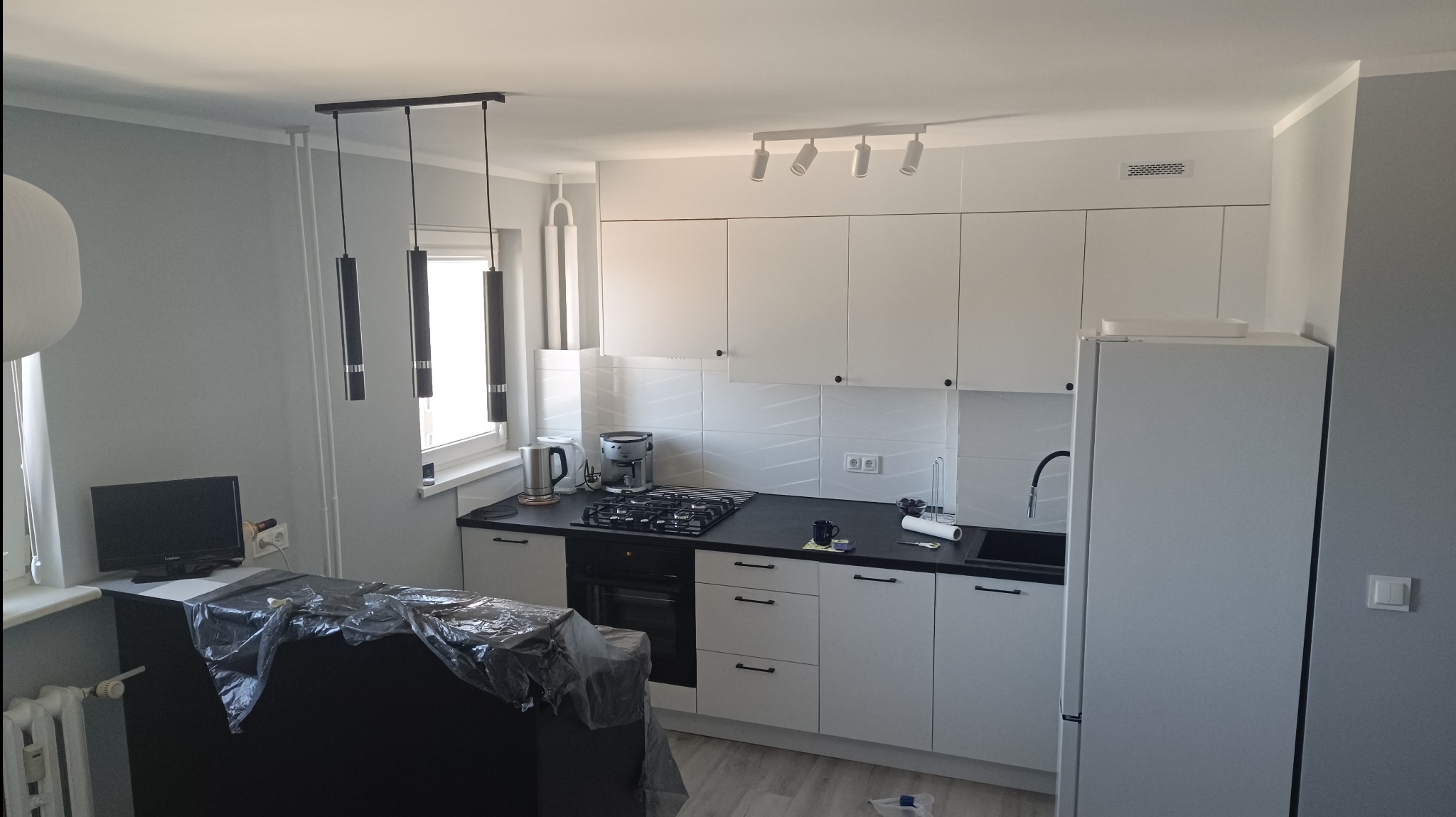 Nowoczesna, jasna kuchnia w szaro-białej tonacji z czarnym blatem, wyposażona w białe szafki z minimalistycznymi uchwytami, czarną płytę grzewczą i piekarnik oraz trzy cylindryczne lampy wiszące...