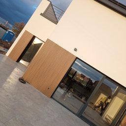 S.perfect-tech Paweł Sperczyński - Nowoczesny dom z jasną elewacją i drewnianymi panelami, widoczny pracownik na drabinie przez okno, w tle pochmurne niebo i przenośna toaleta.