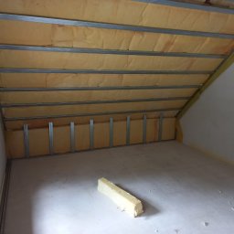 S.perfect-tech Paweł Sperczyński - Poddasze w trakcie ocieplania: wełna mineralna umieszczona między metalowymi profilami, fragment materiału izolacyjnego leżący na podłodze, widok na okno.