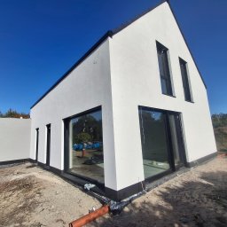 S.perfect-tech Paweł Sperczyński - Nowoczesny dom z białą elewacją i dużymi oknami, widok z zewnątrz na tle błękitnego nieba, teren wokół domu w trakcie prac wykończeniowych, widoczna rura odprowadzająca wodę.