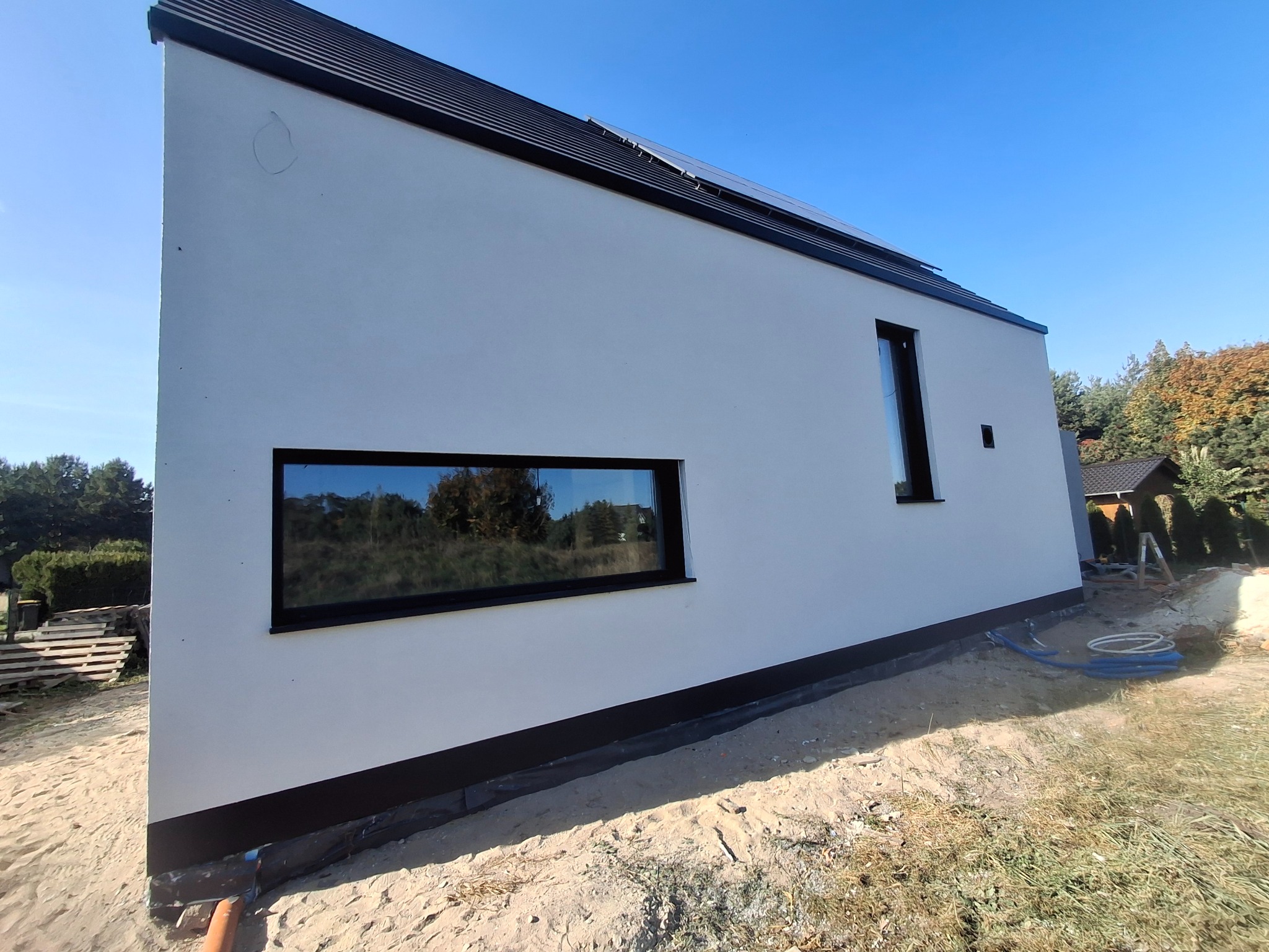 Nowoczesny dom z białą elewacją i czarnymi ramami okiennymi, widok z boku na tle błękitnego nieba i jesiennych drzew, z widocznymi elementami instalacji na gruncie.