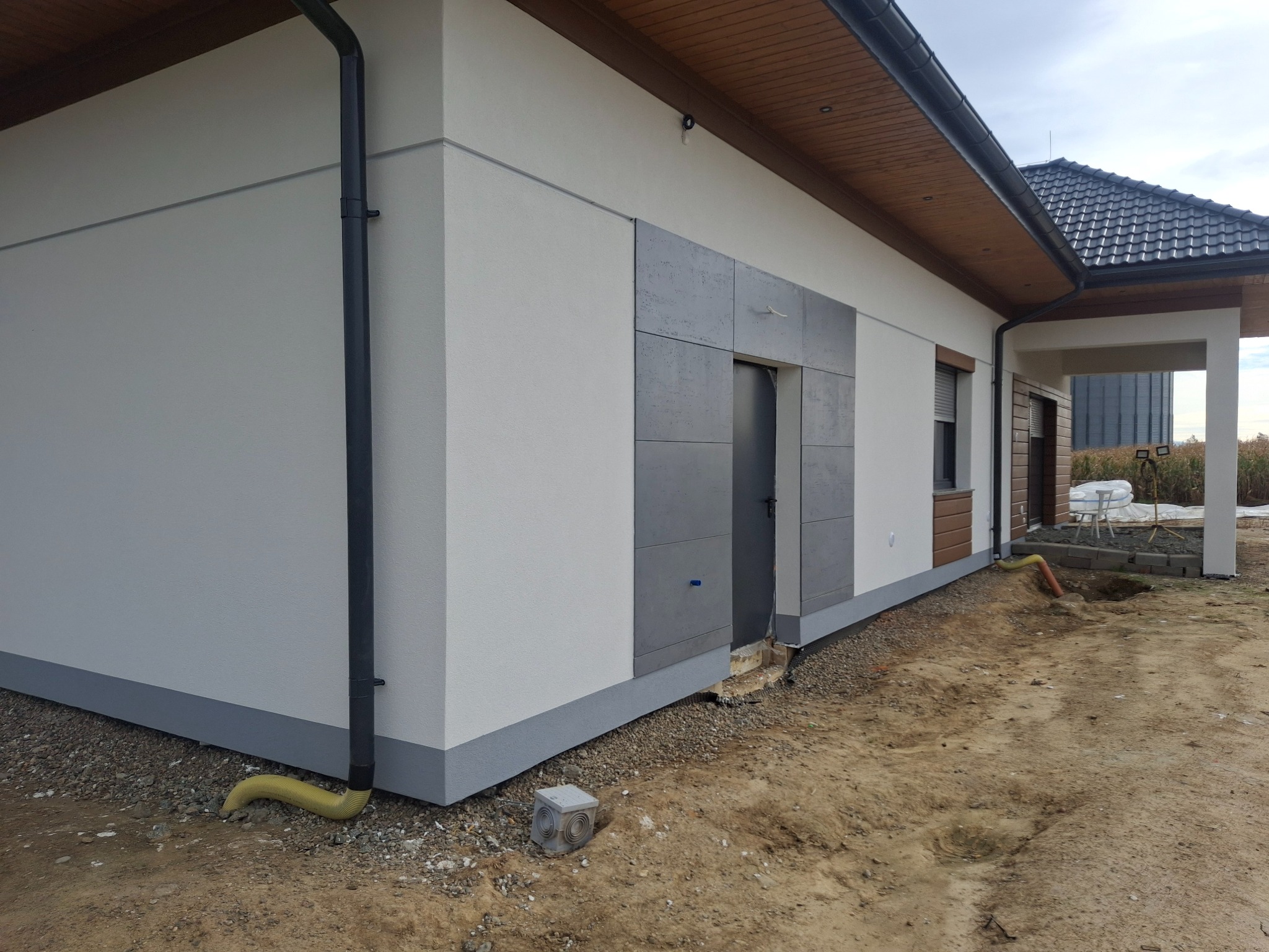 Elewacja domu jednorodzinnego z tynkiem w kolorze złamanej bieli i szarymi panelami dekoracyjnymi wokół drzwi bocznych, widoczna rynna spustowa i fragment terenu przed budynkiem z żółtym wężem...