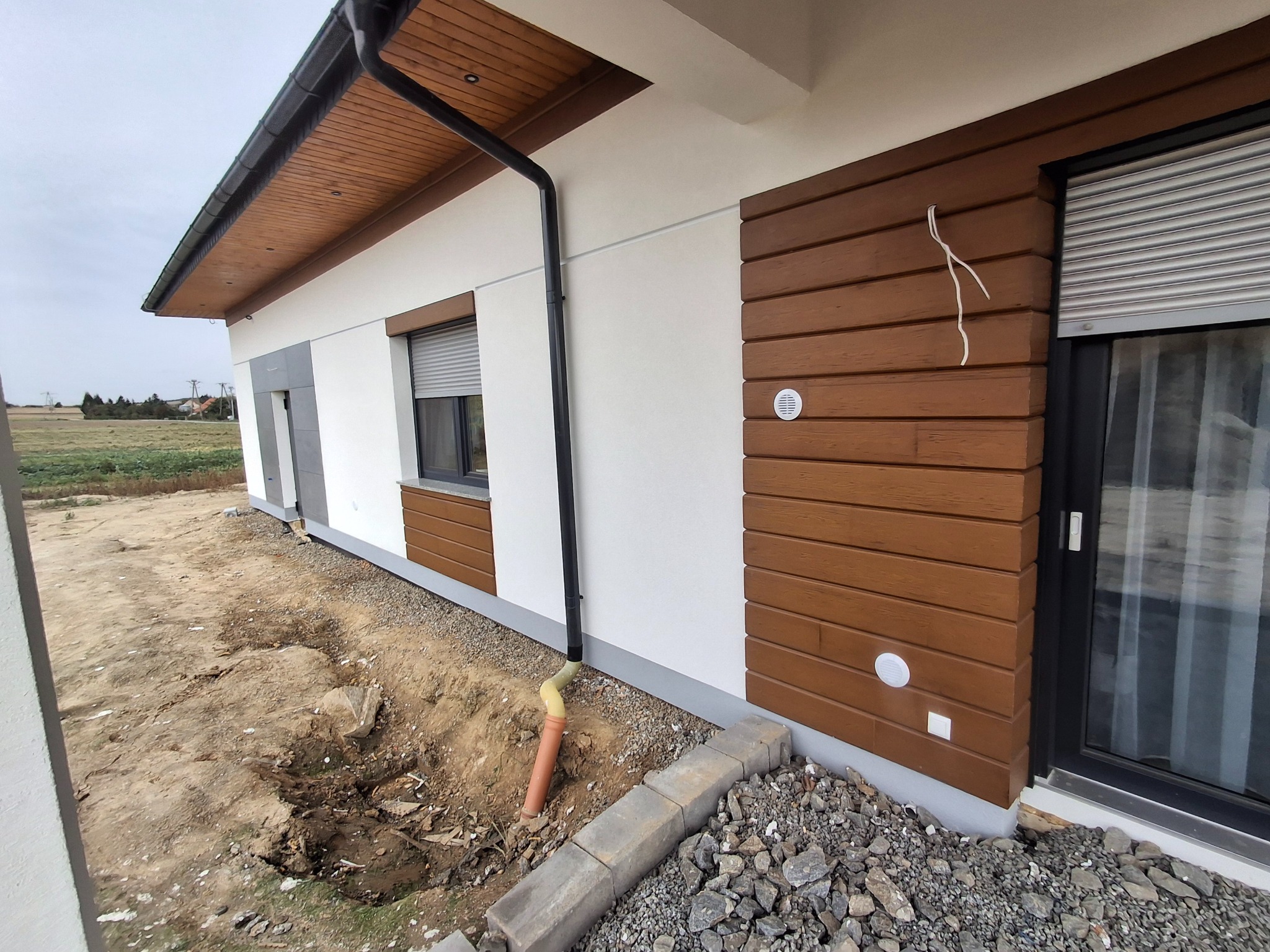 Nowoczesny dom z elewacją w kolorze białym i panelami drewnopodobnymi, widoczne elementy wentylacji, rynna spustowa i częściowo zagospodarowany teren wokół budynku.