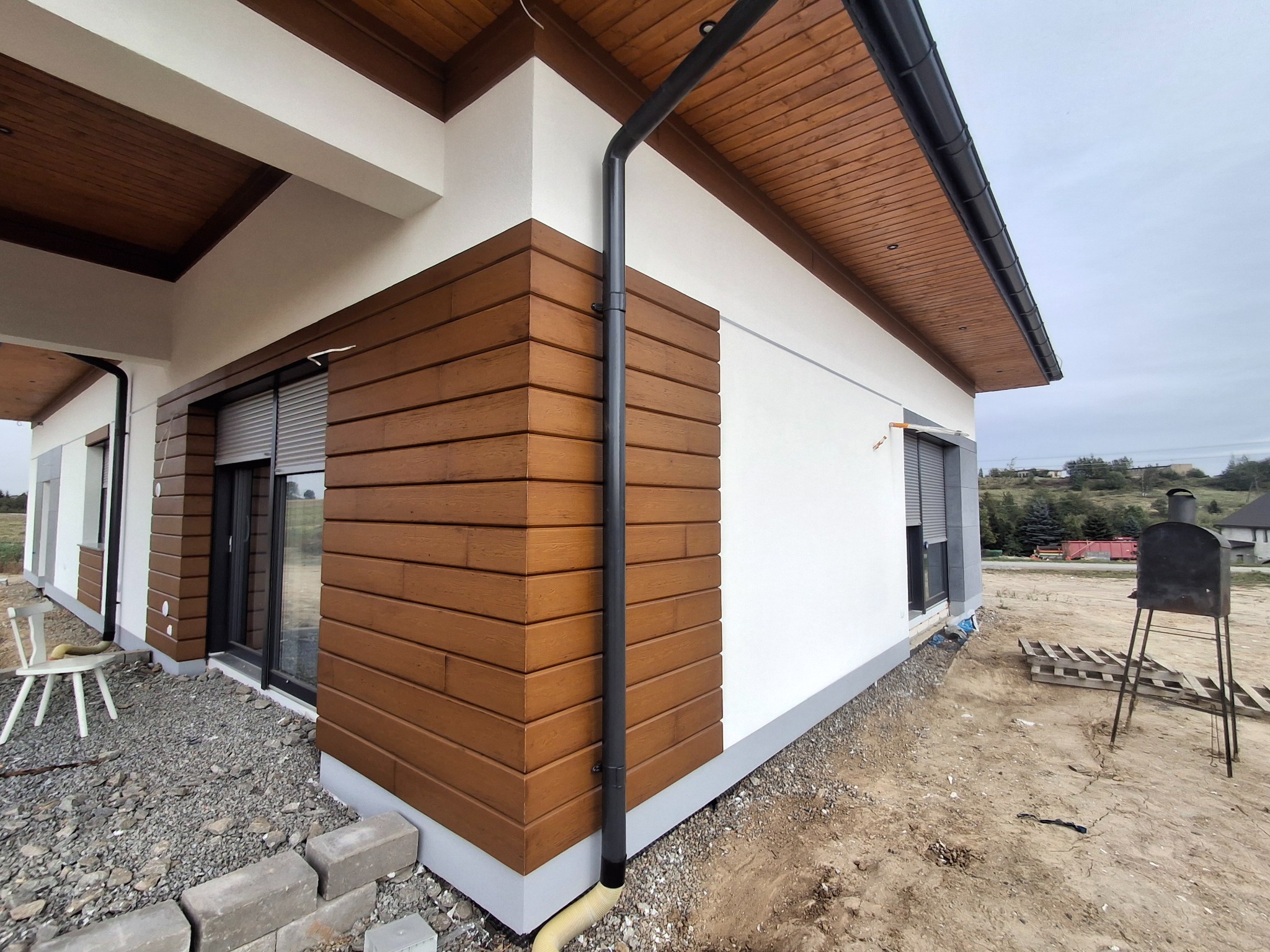 Fragment elewacji domu jednorodzinnego z drewnianą okładziną i oknami z roletami zewnętrznymi, widoczny system rynnowy, teren wokół domu w trakcie prac wykończeniowych.