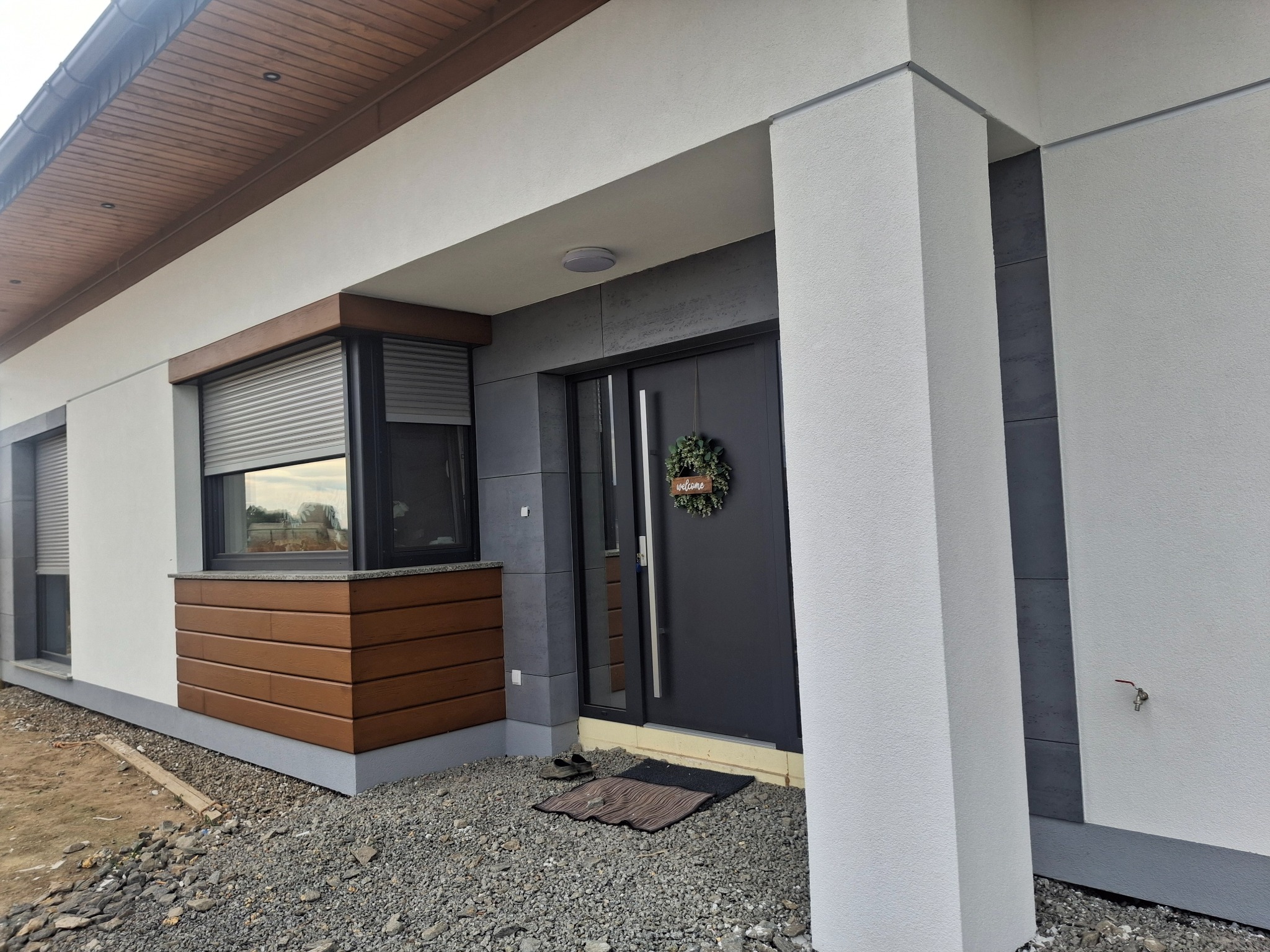 Nowoczesny dom jednorodzinny po renowacji elewacji z elementami dekoracyjnymi z drewna, szarą stolarką okienną z roletami zewnętrznymi i ciemnymi panelami elewacyjnymi, widoczne wykończenie wokół...