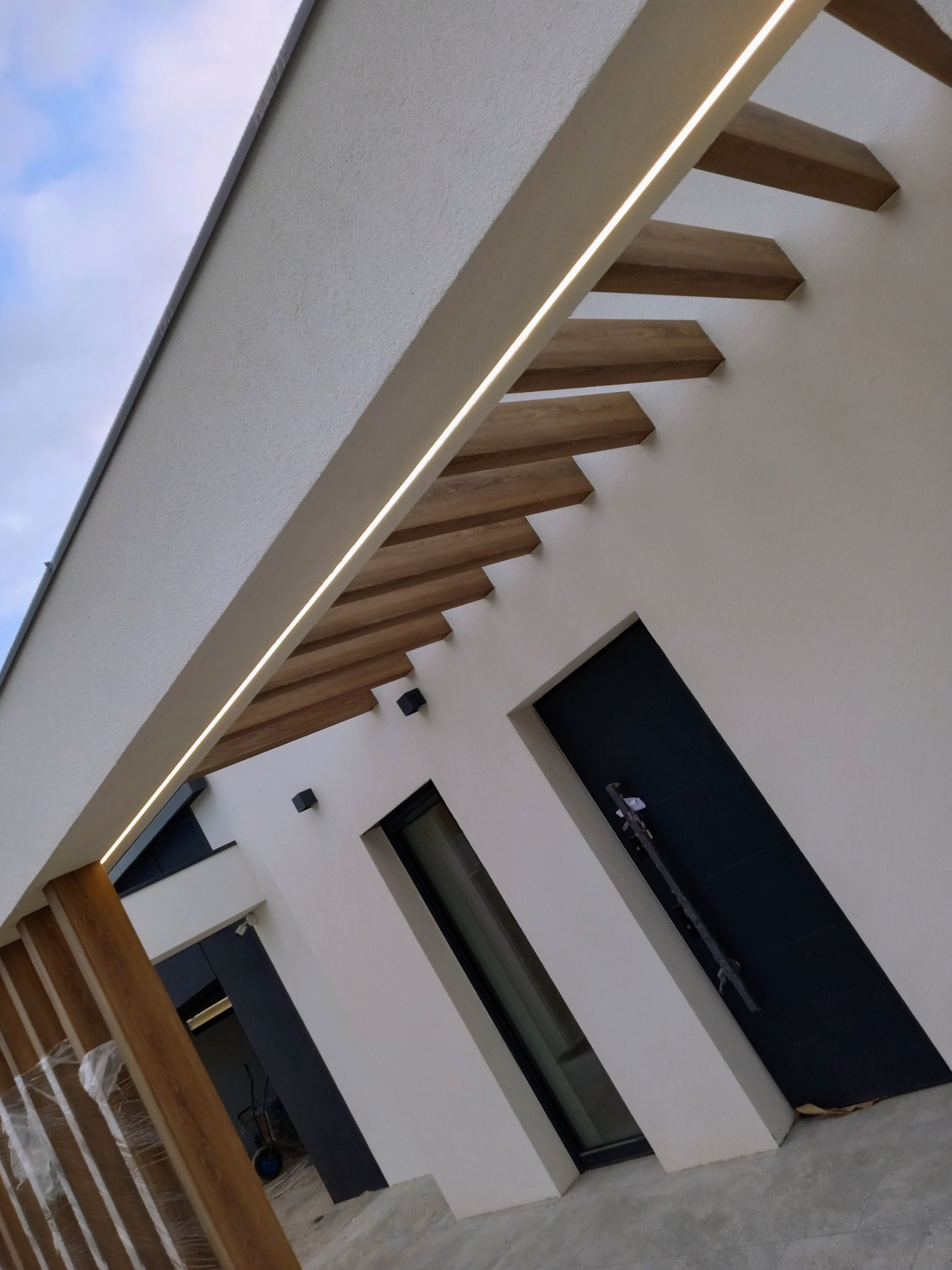 Nowoczesna elewacja domu z oświetleniem LED pod drewnianymi elementami dekoracyjnymi, widoczne okna i drzwi w ciemnej ramie, ujęcie z dołu.