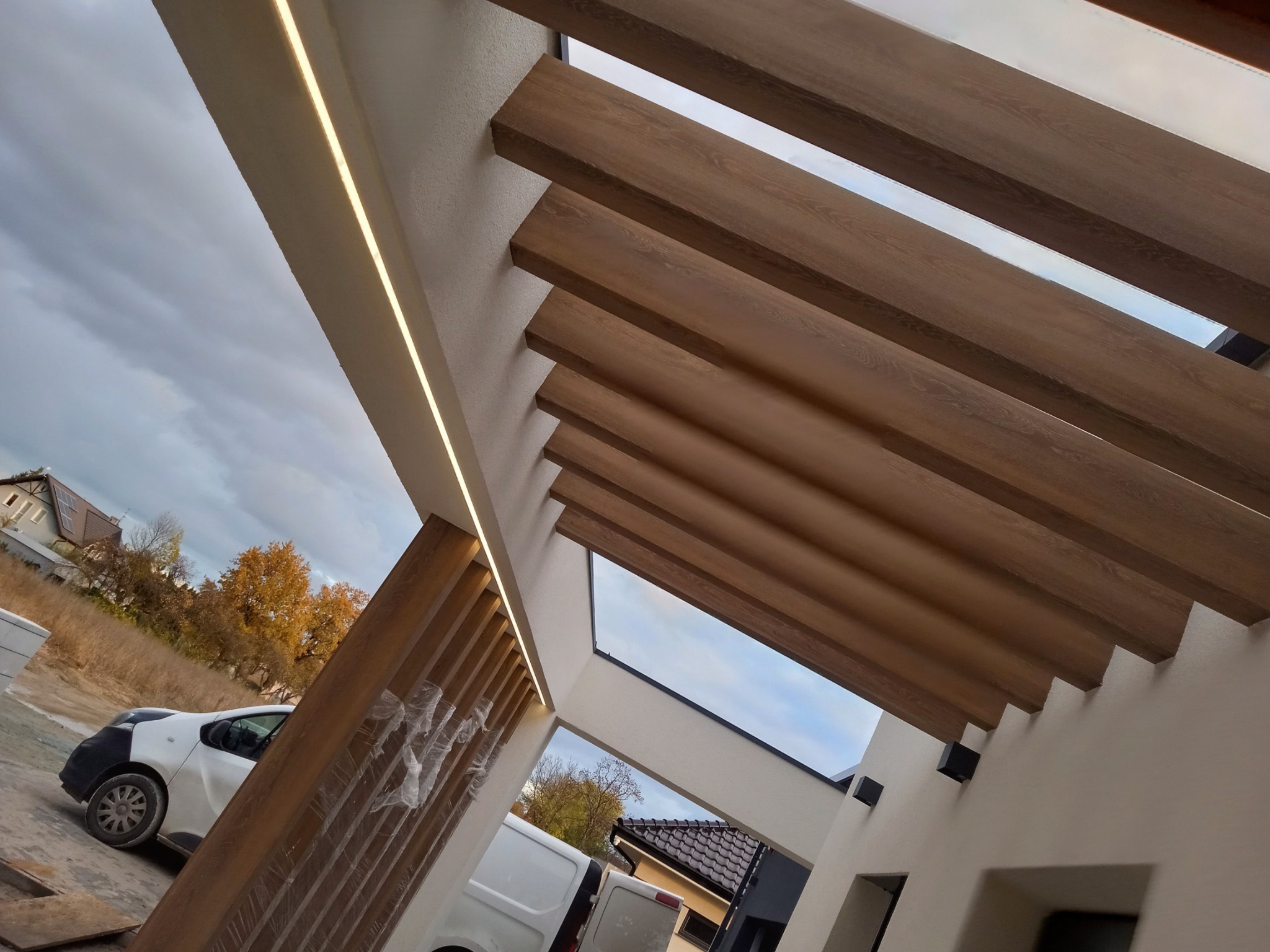 Nowoczesna pergola z drewnianymi belkami i oświetleniem LED w elewacji, widok z dołu na tle pochmurnego nieba i zabudowań.