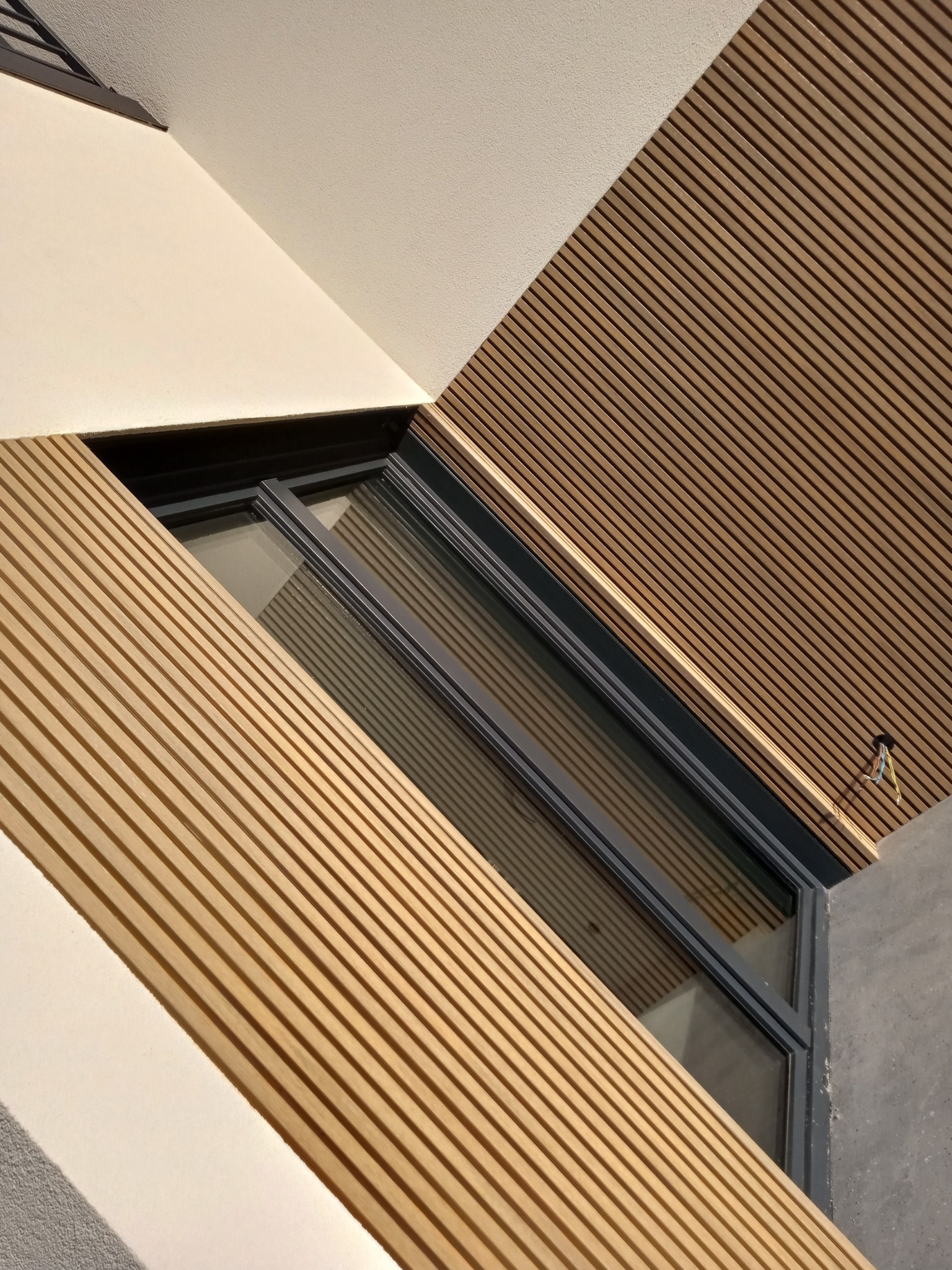 Nowoczesna elewacja budynku z drewnianymi panelami lamelowymi, kremowym tynkiem i oknem z grafitową ramą, widok z dołu.
