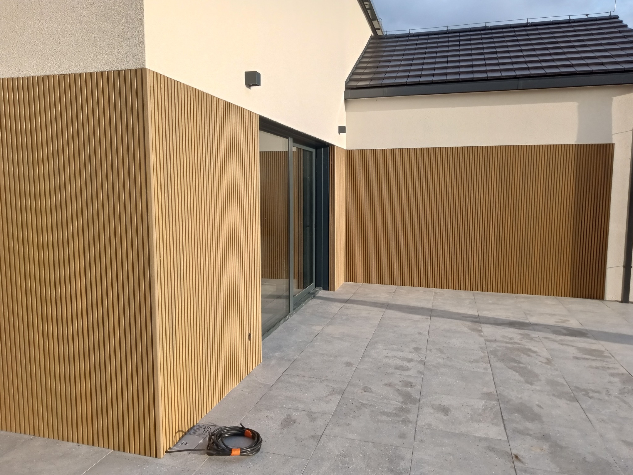 Nowoczesna elewacja budynku z pionowymi panelami w kolorze drewna, połączona z jasnym tynkiem i szarymi płytami chodnikowymi, widoczne czarne lampy elewacyjne i fragment dachu.