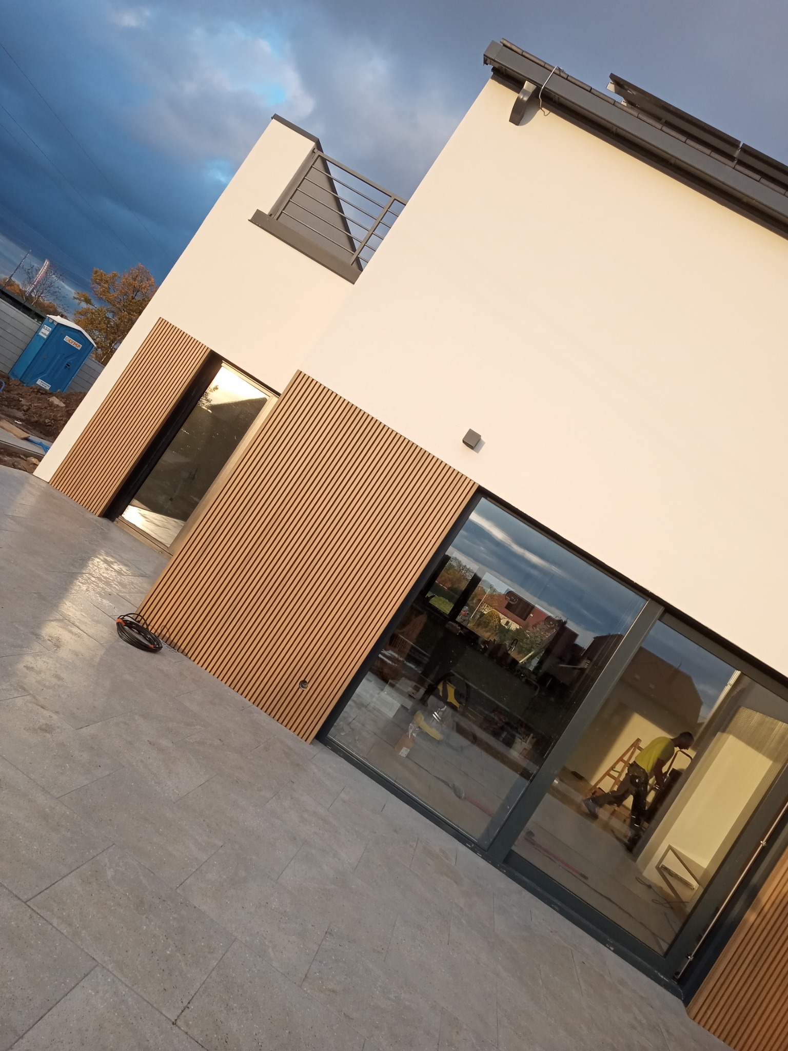 Nowoczesny dom z jasną elewacją i drewnianymi panelami, widoczny pracownik na drabinie przez okno, w tle pochmurne niebo i przenośna toaleta.