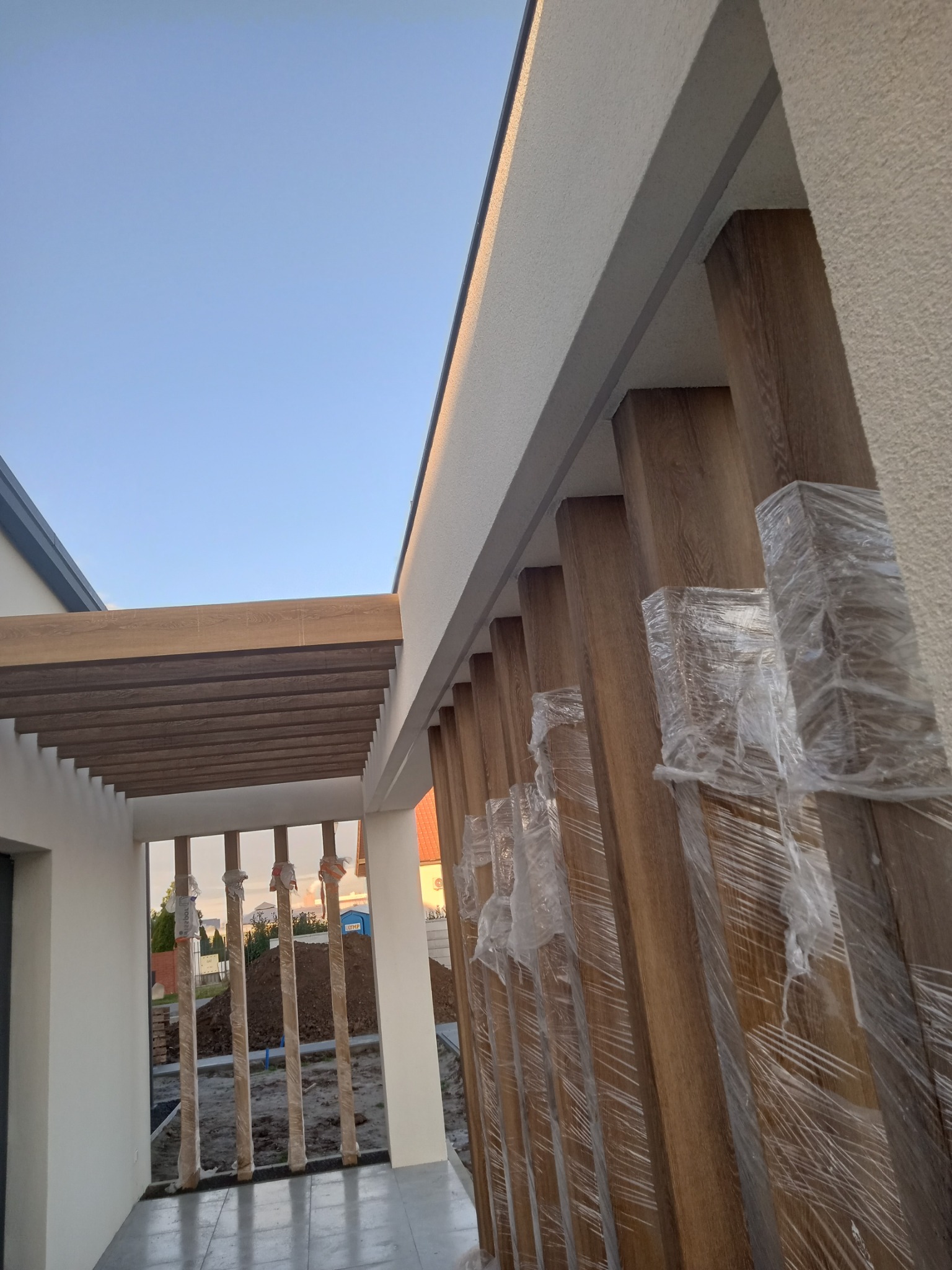 Nowoczesna pergola z drewnopodobnych belek w folii ochronnej, przylegająca do białej ściany budynku w trakcie montażu, widok na przygotowany teren.