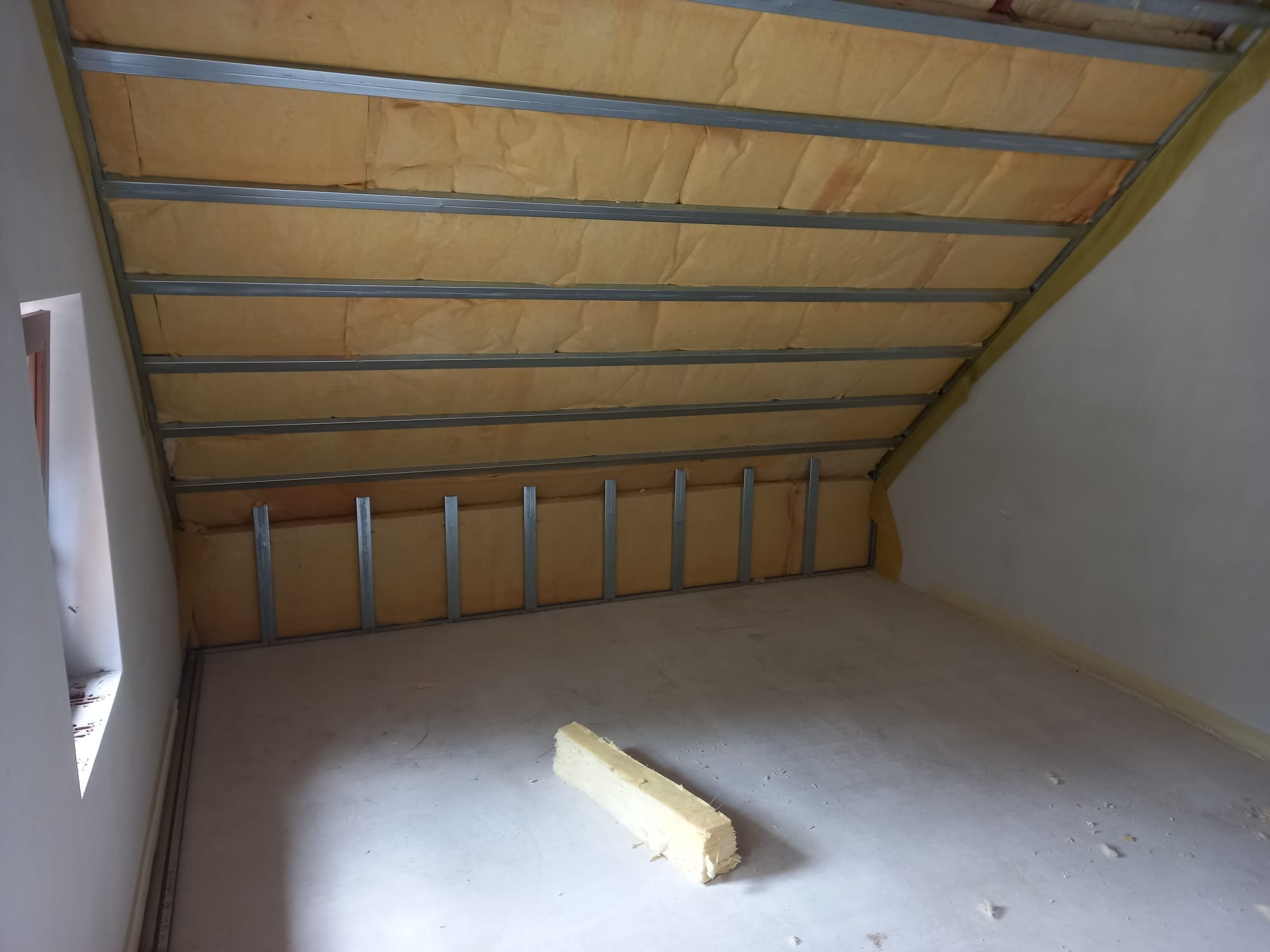 Poddasze w trakcie ocieplania: wełna mineralna umieszczona między metalowymi profilami, fragment materiału izolacyjnego leżący na podłodze, widok na okno.