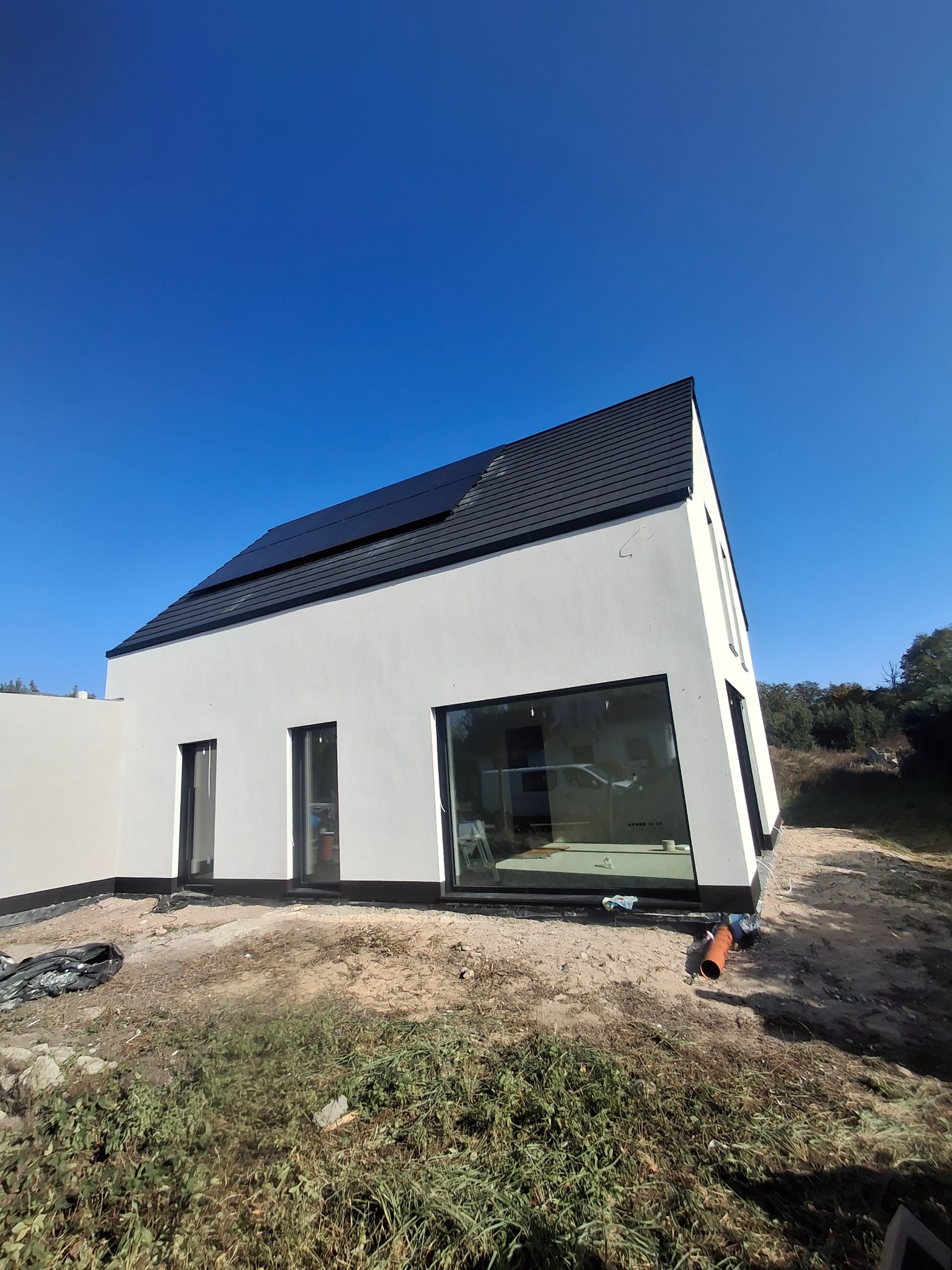 Nowoczesny dom jednorodzinny z białą, gładką elewacją, czarną stolarką okienną i panelami słonecznymi na dachu, na działce w trakcie zagospodarowywania.