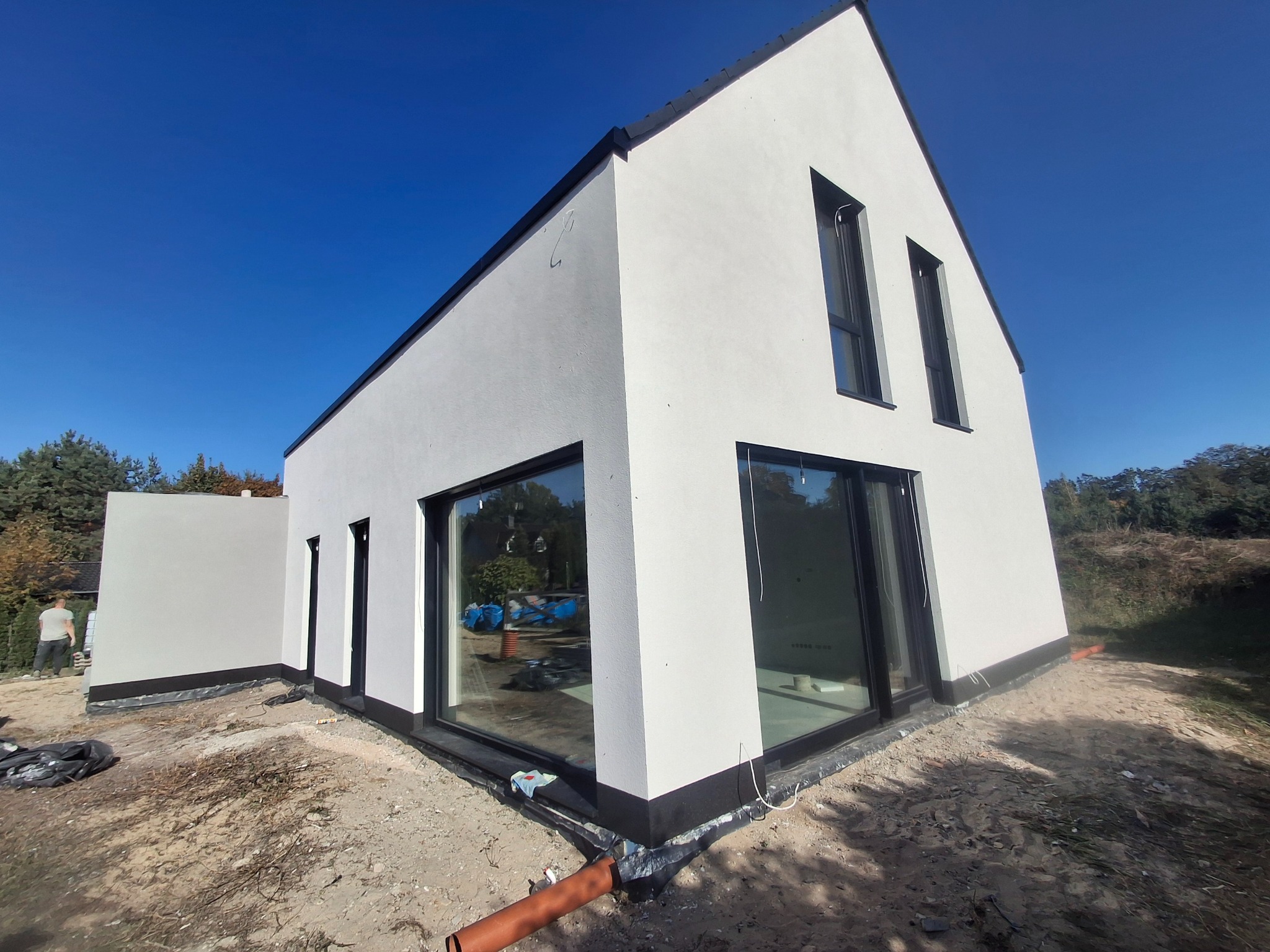 Nowoczesny dom z białą elewacją i dużymi oknami, widok z zewnątrz na tle błękitnego nieba, teren wokół domu w trakcie prac wykończeniowych, widoczna rura odprowadzająca wodę.