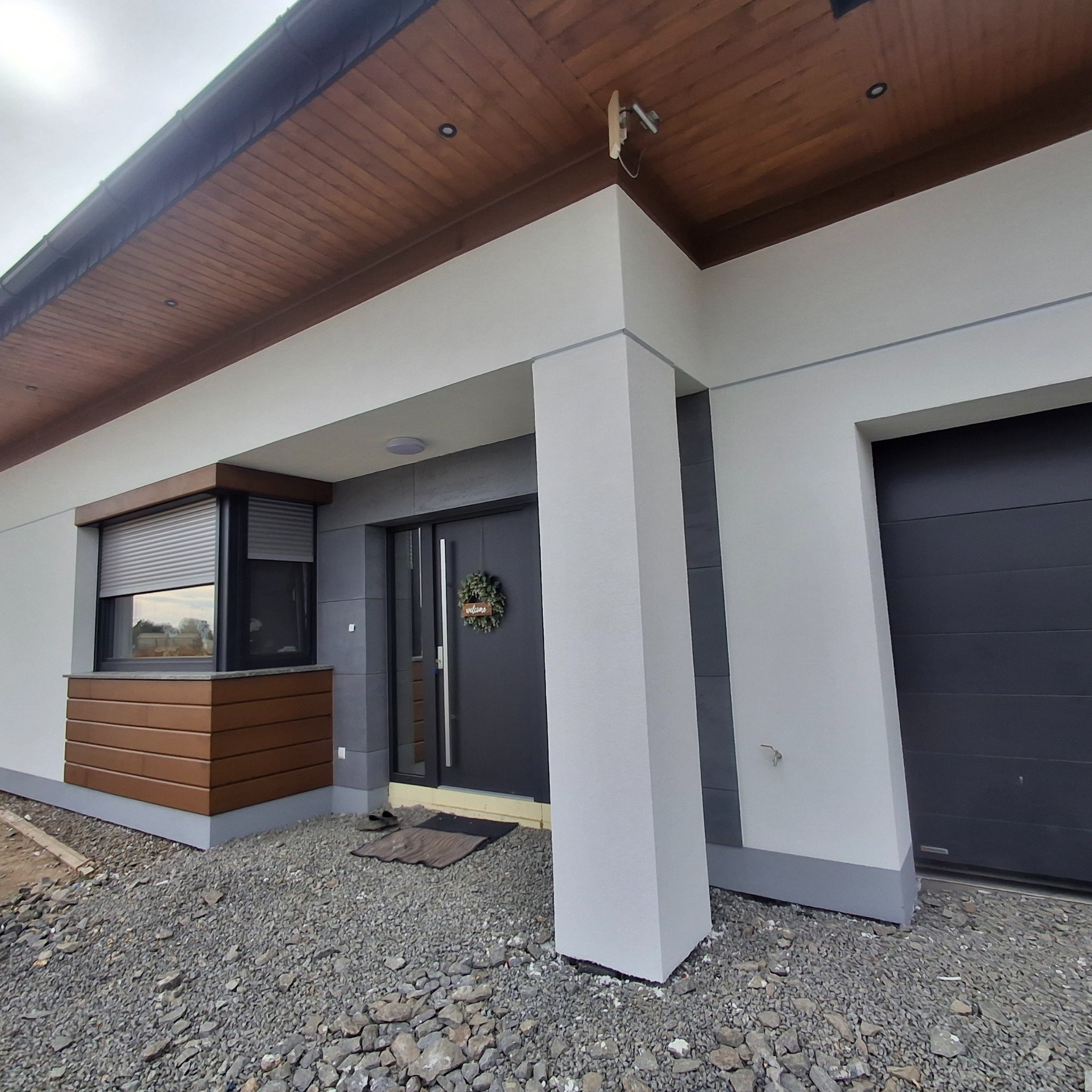 Nowoczesny dom jednorodzinny z szarą elewacją, drewnianym podbitką dachu i roletami zewnętrznymi, widok z poziomu gruntu na wejście z dekoracyjnym wieńcem.