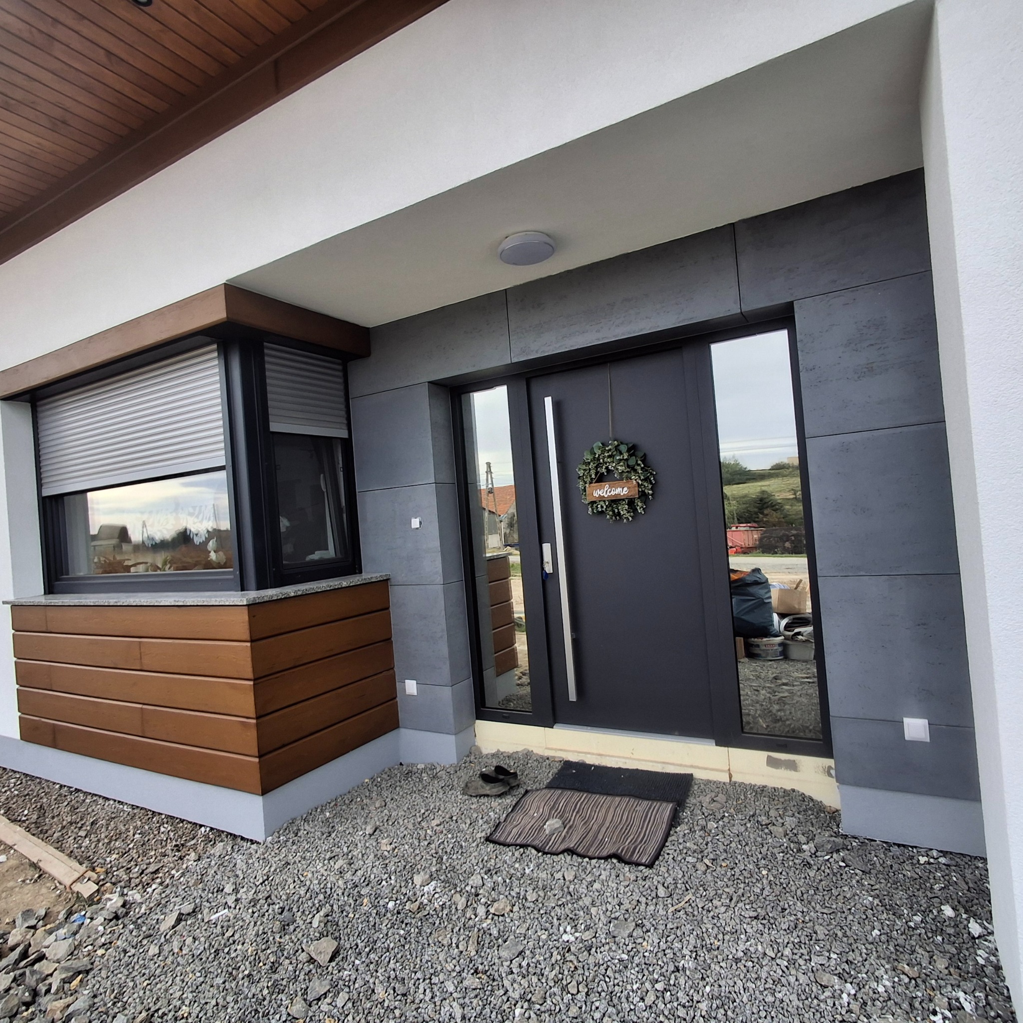 Nowoczesna elewacja domu z szarymi panelami, ciemnymi drzwiami wejściowymi z dekoracyjnym wieńcem i oknem z roletami zewnętrznymi, wykończona drewnianymi elementami. Podłoże wysypane drobnym żwirem.