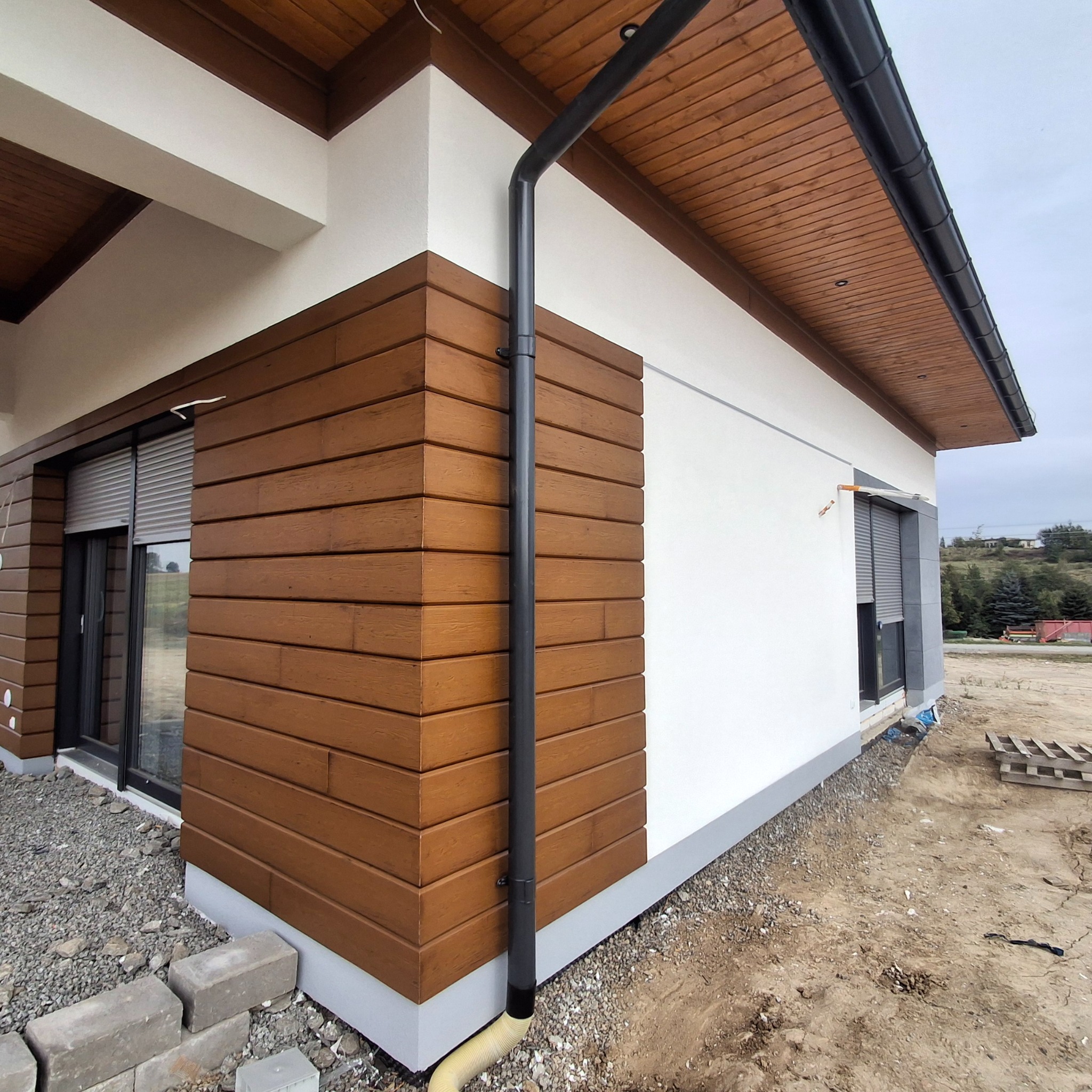 Nowoczesny dom z elewacją w dwóch kolorach: biała ściana i brązowe, poziome panele imitujące drewno, czarna rynna, okno z roletą zewnętrzną, teren wokół domu w trakcie prac wykończeniowych.