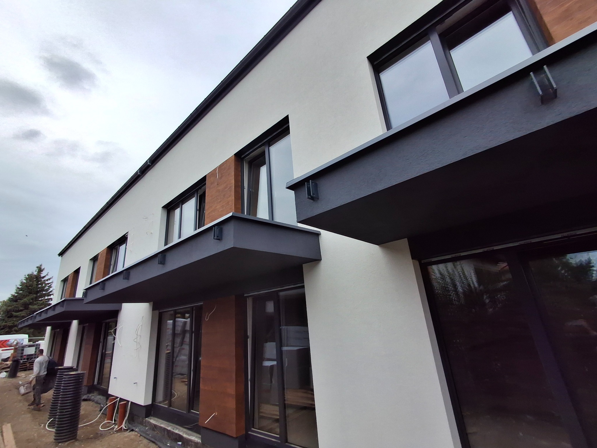 Nowoczesna elewacja budynku szeregowego z drewnianymi akcentami i szarymi nadprożami, widok z dołu, częściowo widoczne prace wykończeniowe.