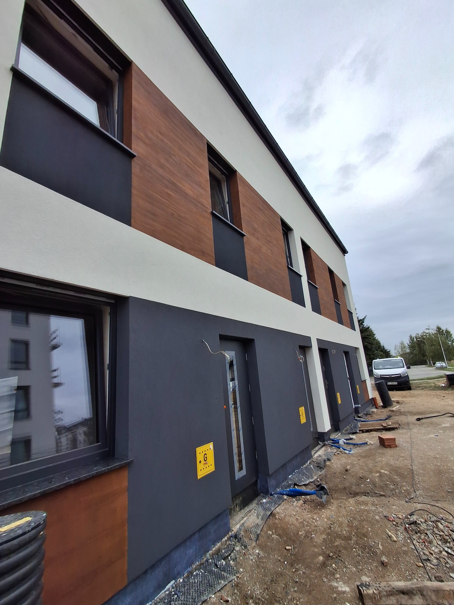 Nowoczesna elewacja budynku z elementami drewnopodobnymi i szarą farbą, widok z dołu, teren budowy w tle. Detal wykończenia.