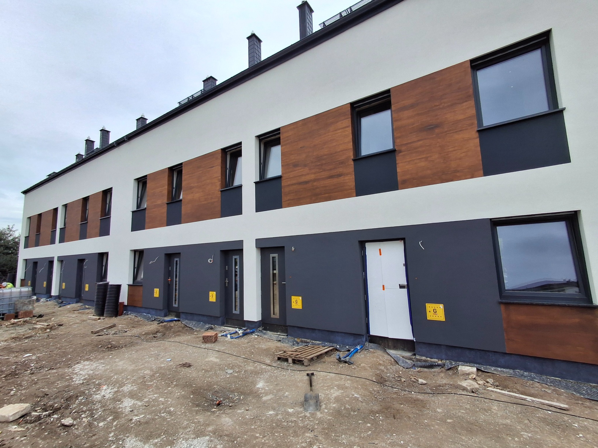 Nowoczesny budynek szeregowy z elewacją w odcieniach bieli, szarości i drewna. Widoczne okna, drzwi i fragment placu budowy z ziemią i materiałami.