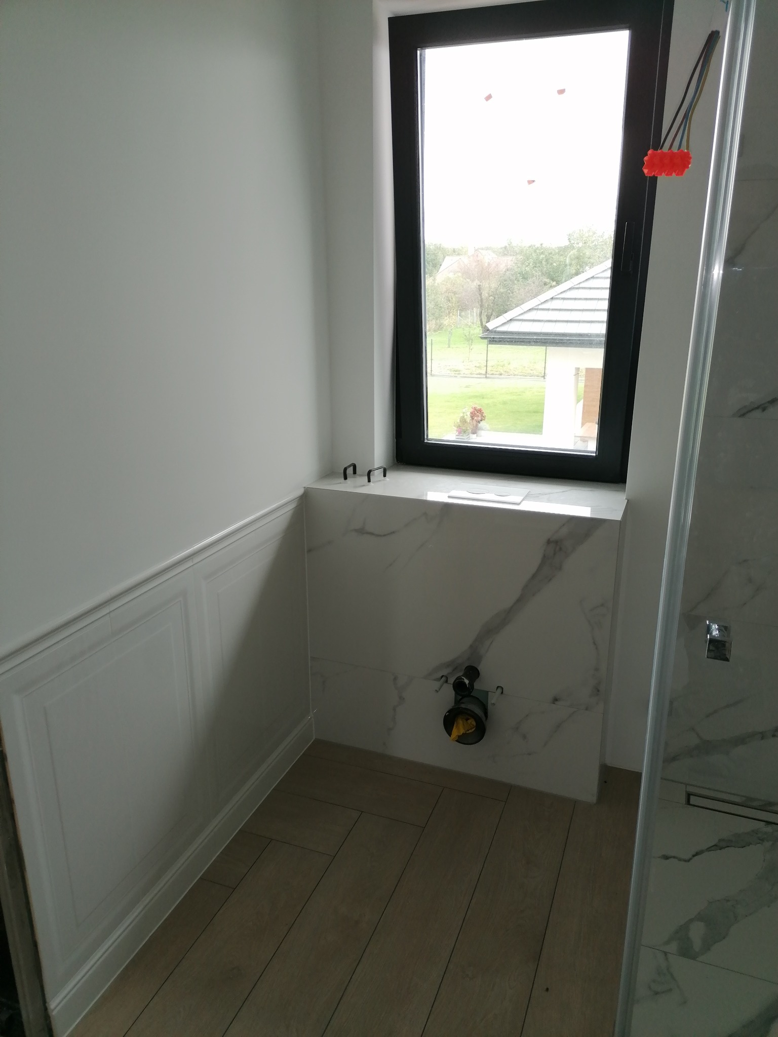 Przygotowanie do montażu stelaża podtynkowego WC w łazience z marmurową okładziną, widok na okno z czarną ramą i fragment ściany z dekoracyjną sztukaterią.