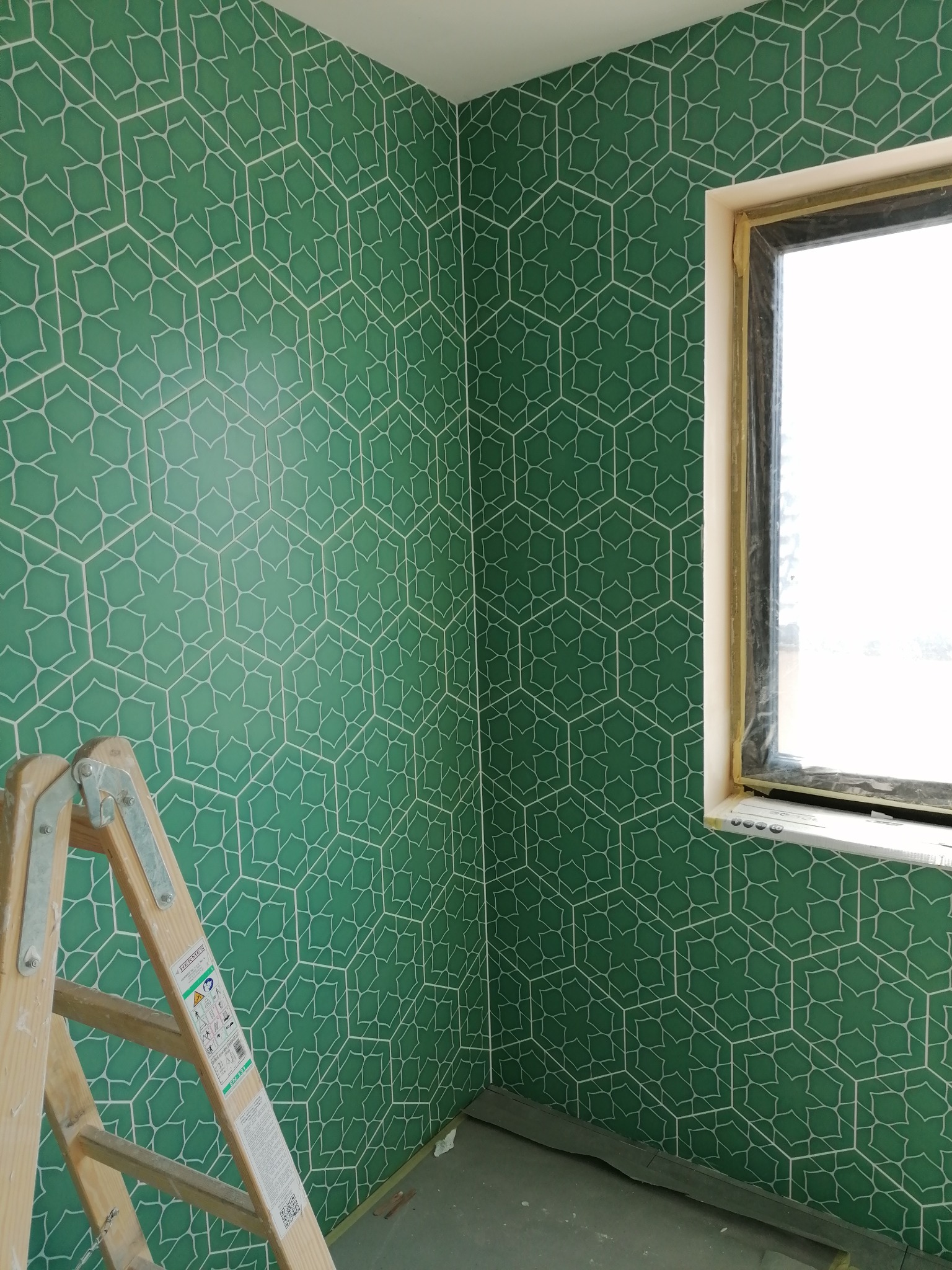 Ściana w rogu pomieszczenia wyłożona zielonymi płytkami z białym geometrycznym wzorem, widoczne okno z czarną ramą i drewniana drabina oparta o ścianę