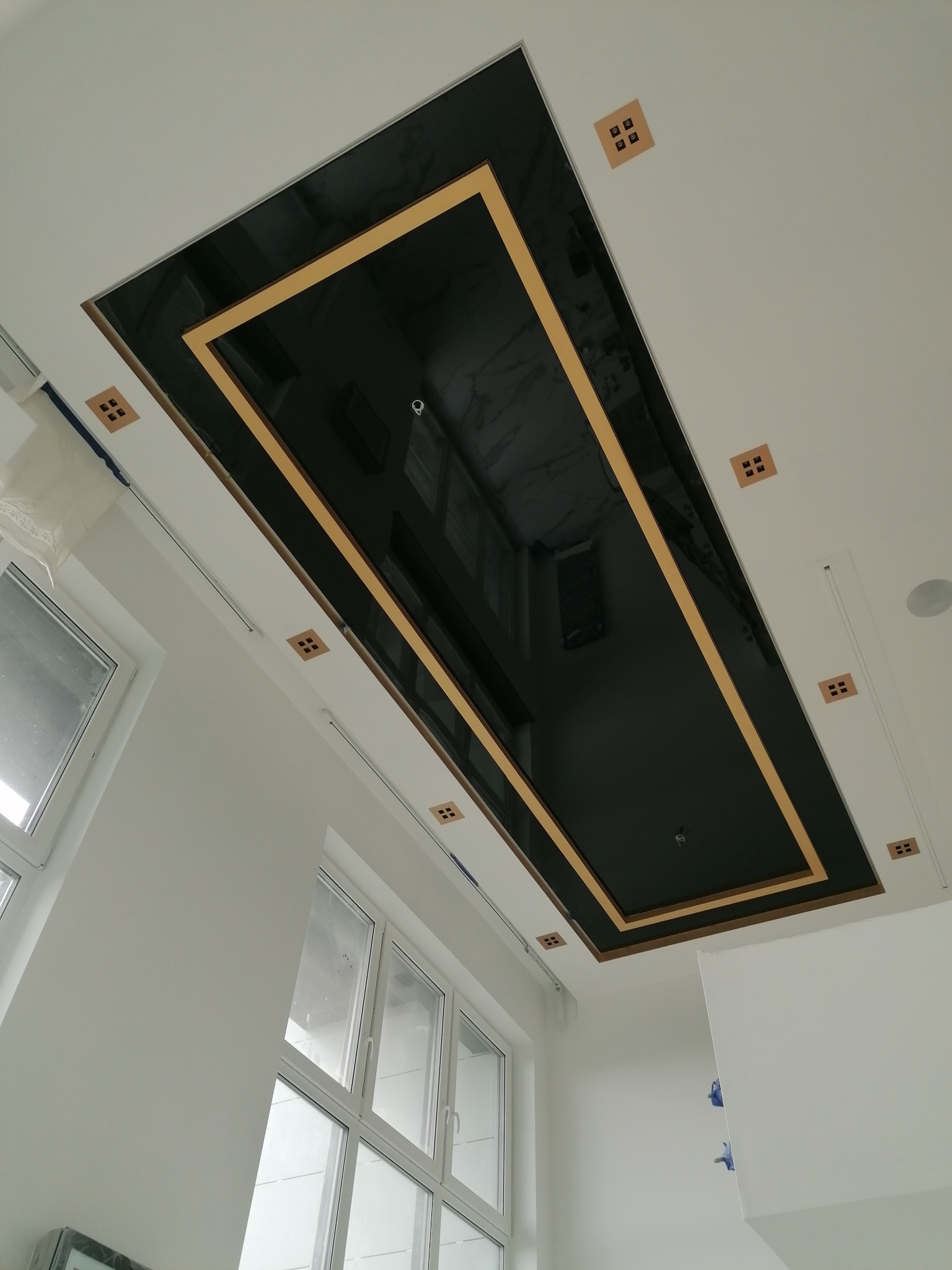 Nowoczesny sufit podwieszany z czarną, błyszczącą powierzchnią i złotą ramą, otoczony białymi ścianami i oknami, z ozdobnymi kwadratowymi elementami na suficie.