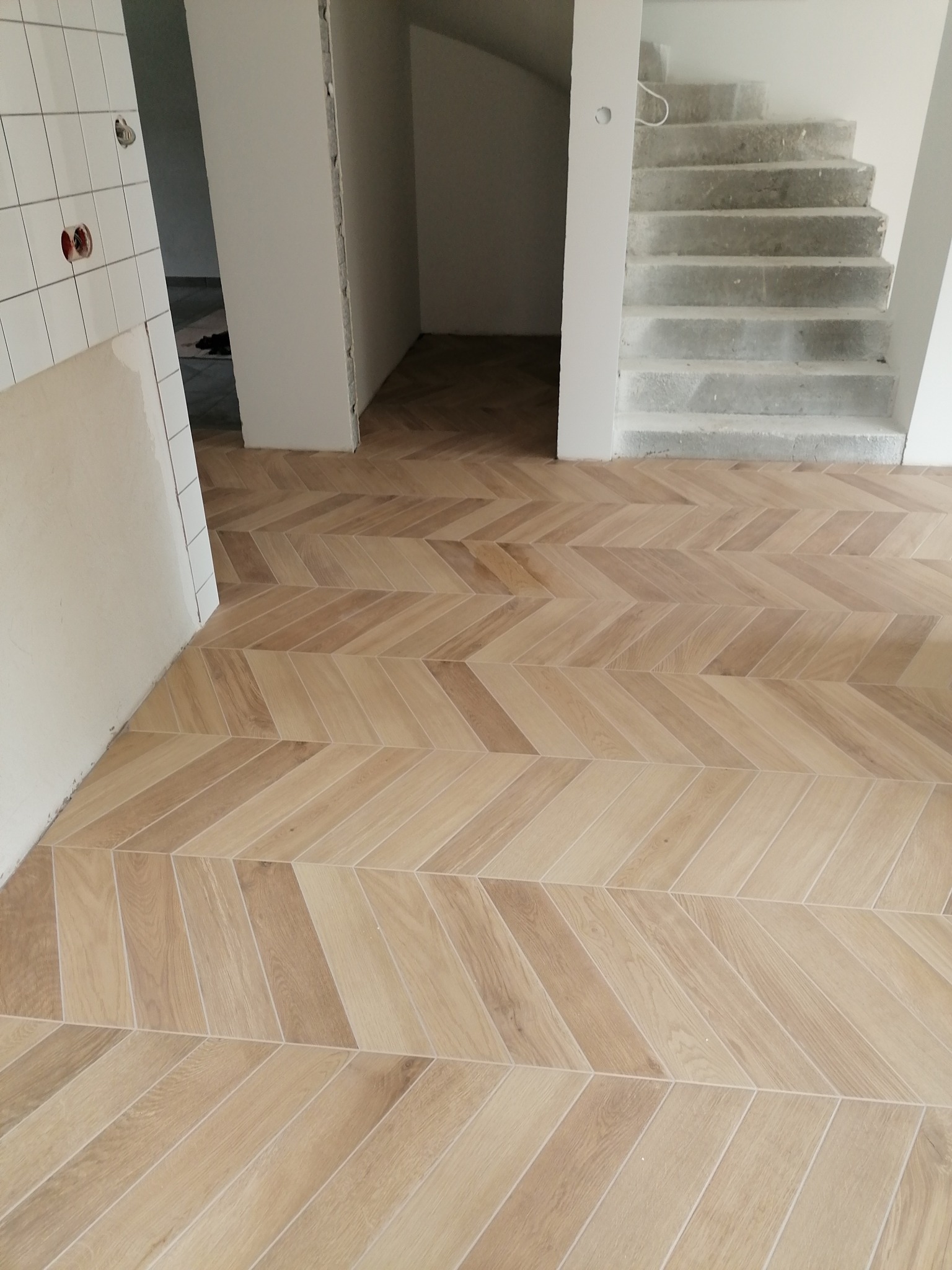 Ułożona w jodełkę podłoga z paneli drewnopodobnych, widok na surowe betonowe schody i fragment ściany z białymi płytkami w trakcie prac wykończeniowych.