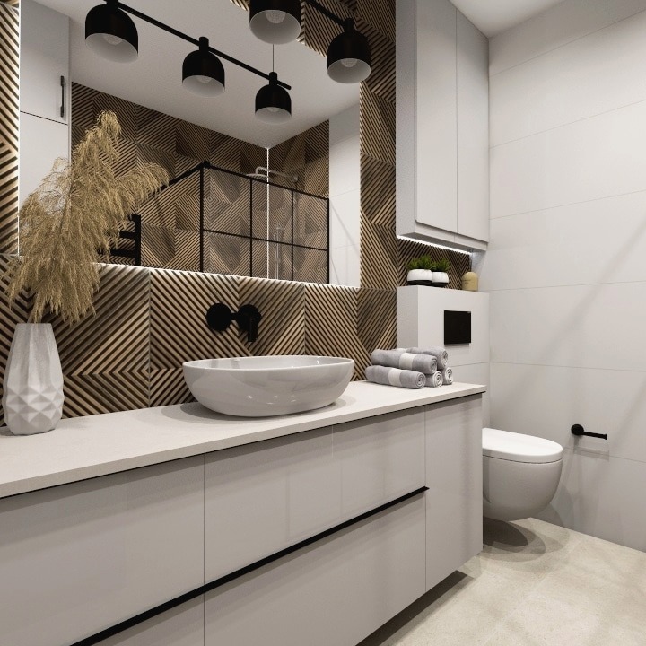 Nowoczesna łazienka z drewnianą mozaiką na ścianie, biała umywalka nablatowa z czarną armaturą, szara szafka pod umywalką z czarnymi uchwytami, dekoracyjna trawa w wazonie, białe płytki i toaleta.