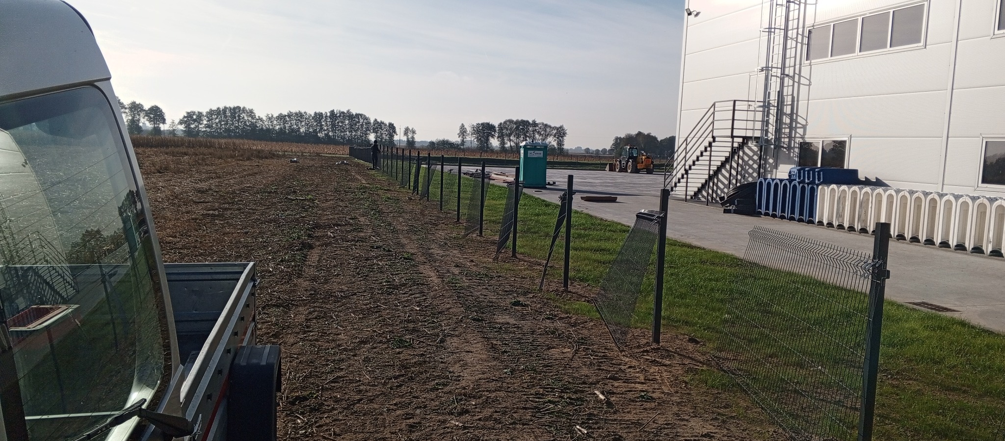 Długi odcinek nowo postawionego ogrodzenia panelowego z metalowej siatki, biegnący wzdłuż pola uprawnego i budynku przemysłowego. Widoczny fragment przyczepy transportowej. W tle traktor...