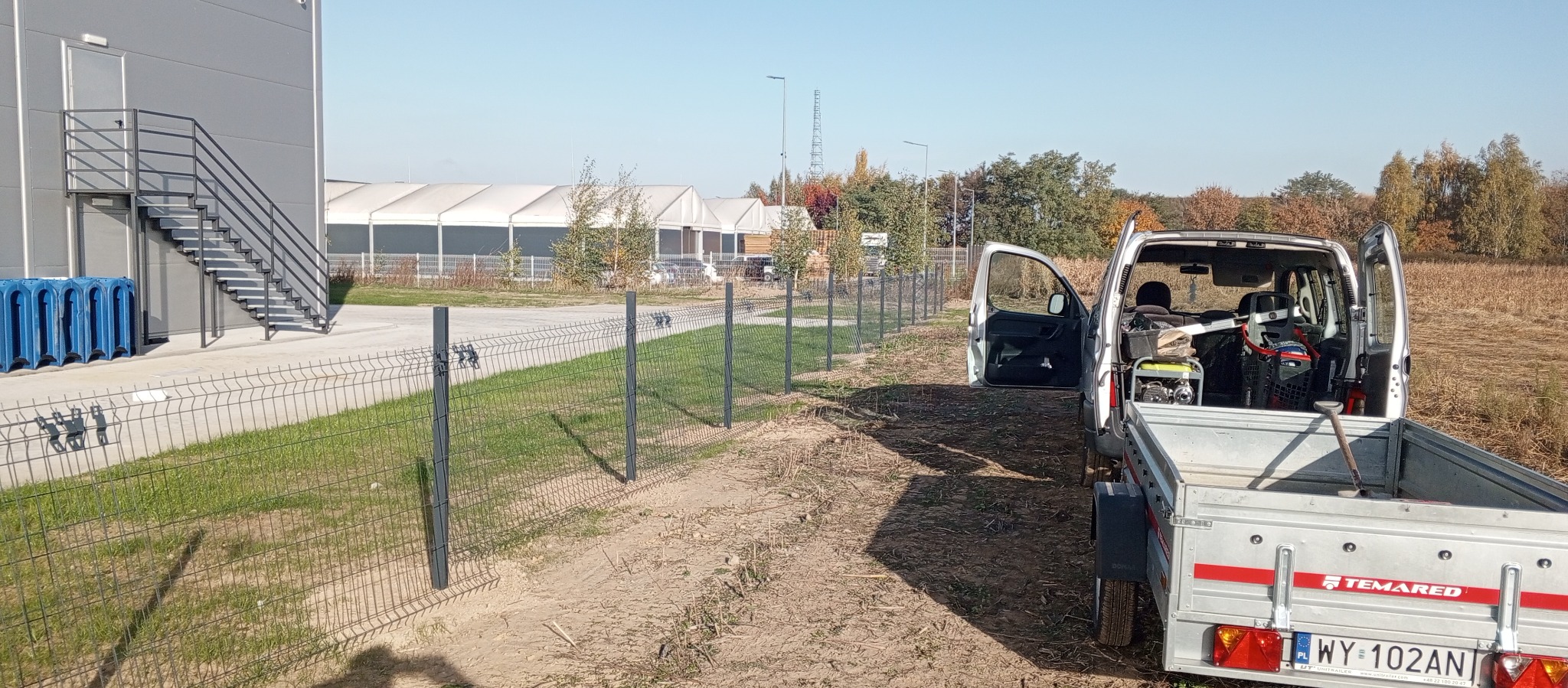 Fragment nowo postawionego, panelowego ogrodzenia z metalowej siatki w szarym kolorze, widoczny samochód dostawczy z otwartymi drzwiami i przyczepką z narzędziami w tle, teren trawiasty i budynek...