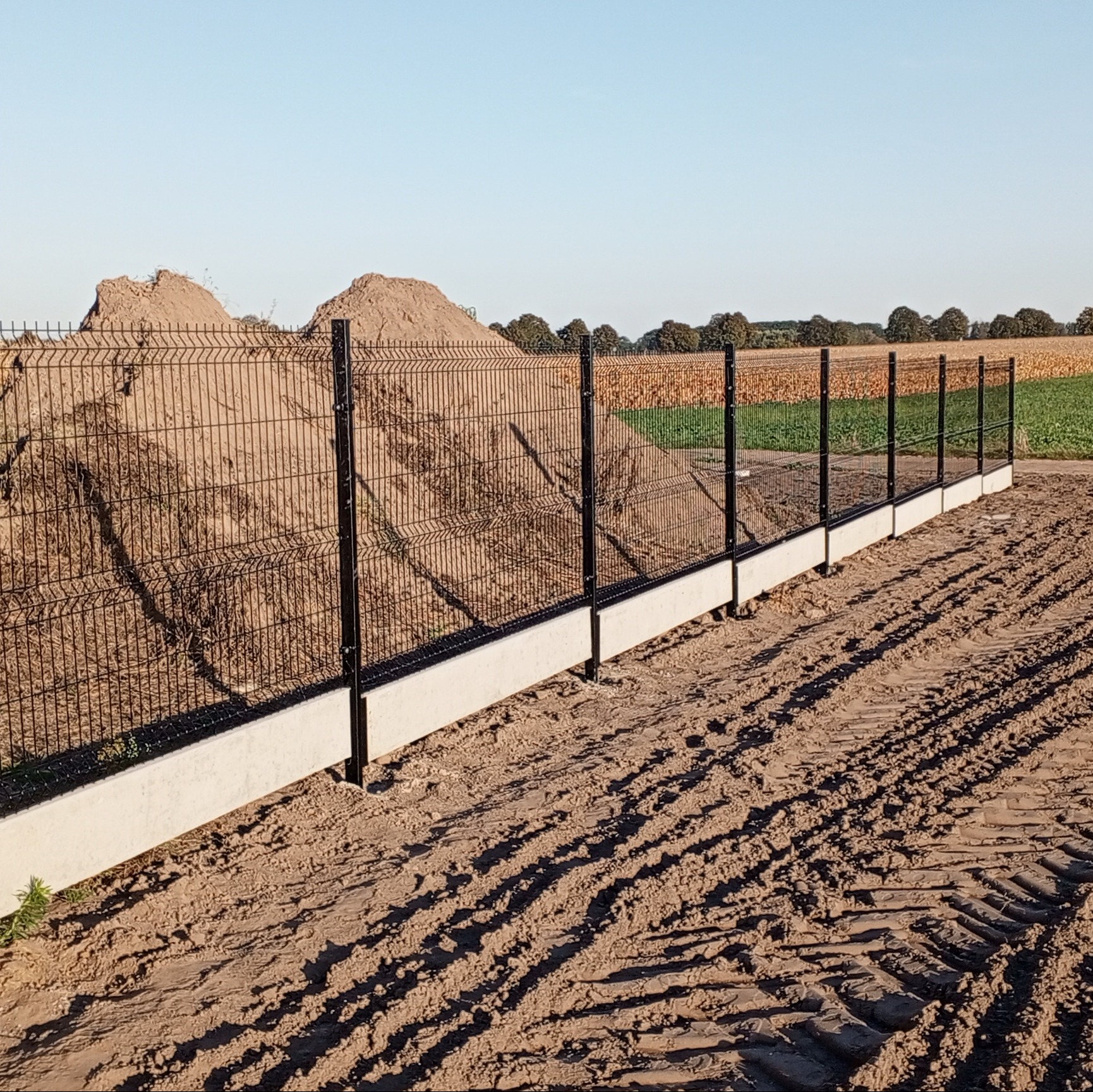 Nowoczesne ogrodzenie panelowe z czarnymi słupkami i betonową podmurówką na nierównym terenie z pryzmą piasku w tle, widoczne ślady opon.