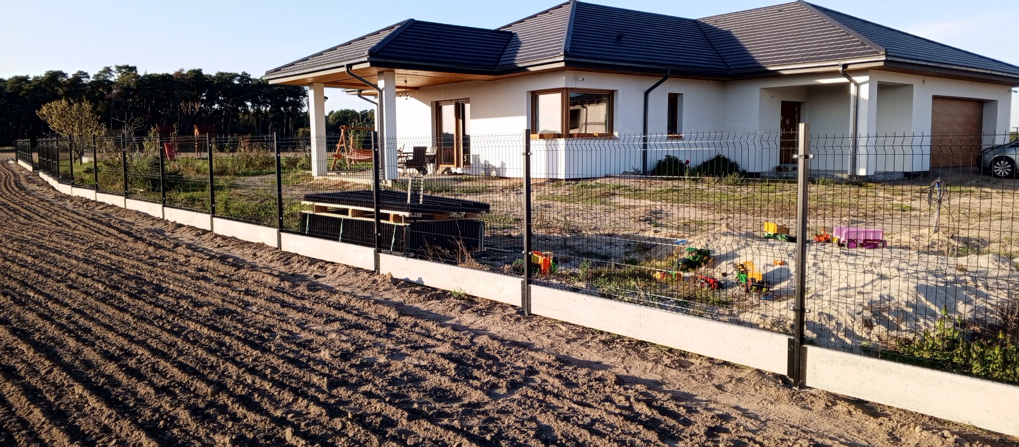 Nowoczesne ogrodzenie panelowe z podmurówką betonową, widok na dom z garażem i plac zabaw z zabawkami dziecięcymi za ogrodzeniem.